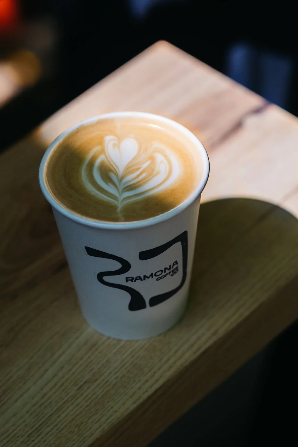A disposable cup with a coffee drink featuring latte art in a heart shape, on a wooden surface inside a coffee shop.