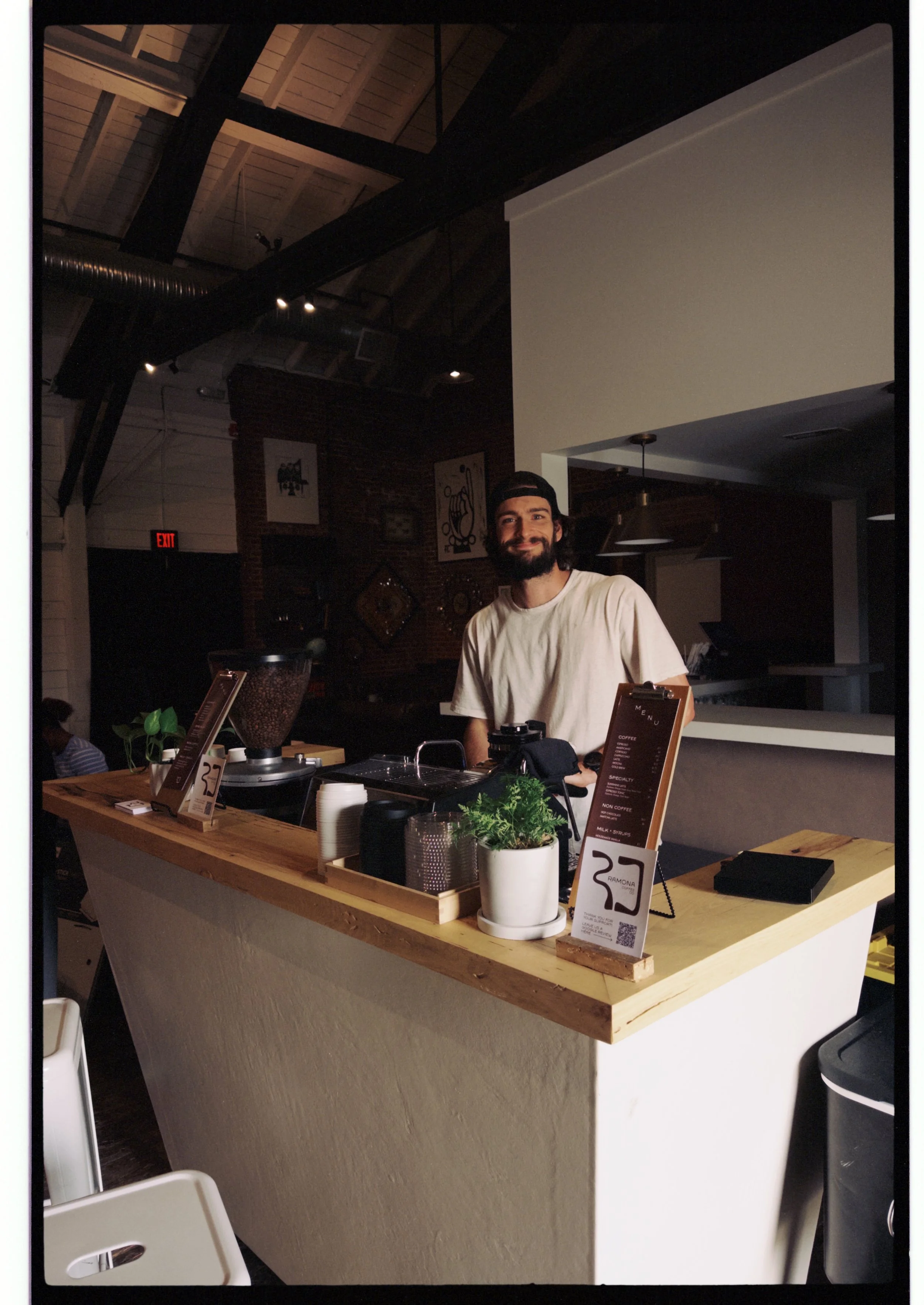 A barista stands behind a wooden counter at a coffee shop, smiling. The counter has potted plants, a menu sign, and coffee-making equipment. The background features a brick wall with art and dark ceiling with exposed beams and ductwork.