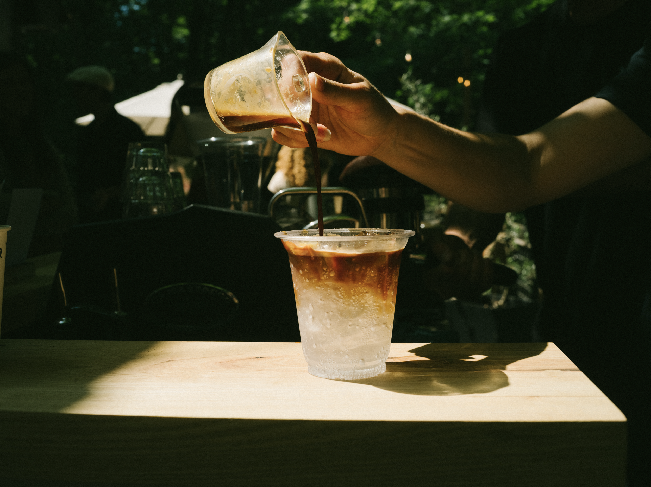 A hand pouring dark coffee from a glass container into a clear plastic cup on a wooden table outdoors, with sunlight and trees in the background.