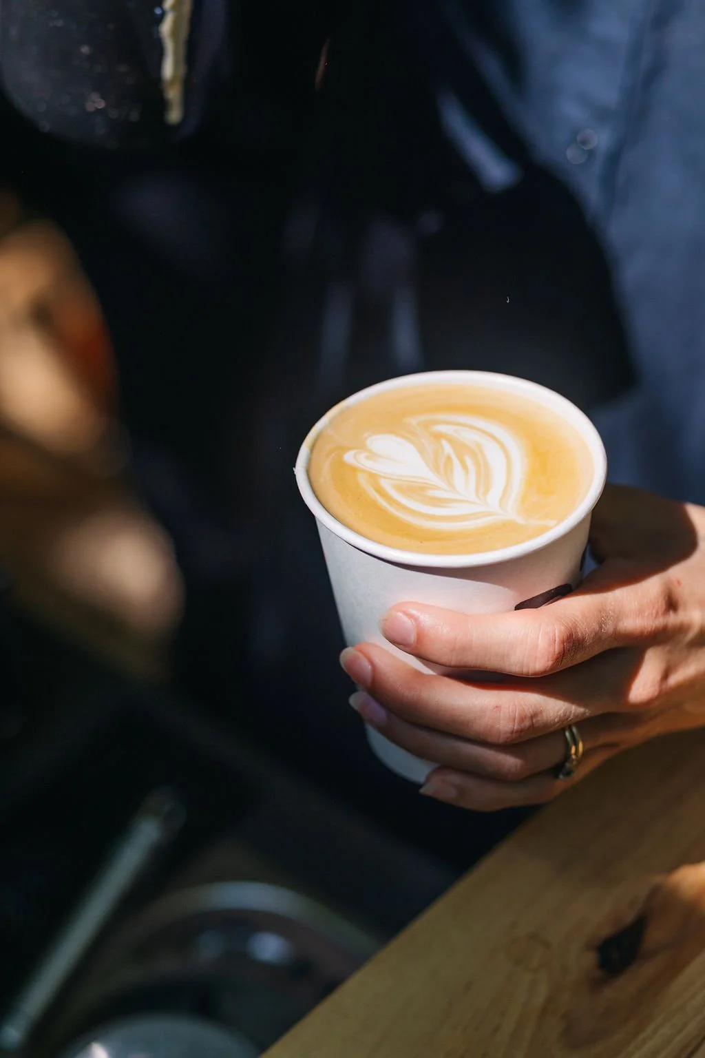 A person holding a paper cup of latte with latte art in the shape of a heart and leaves.