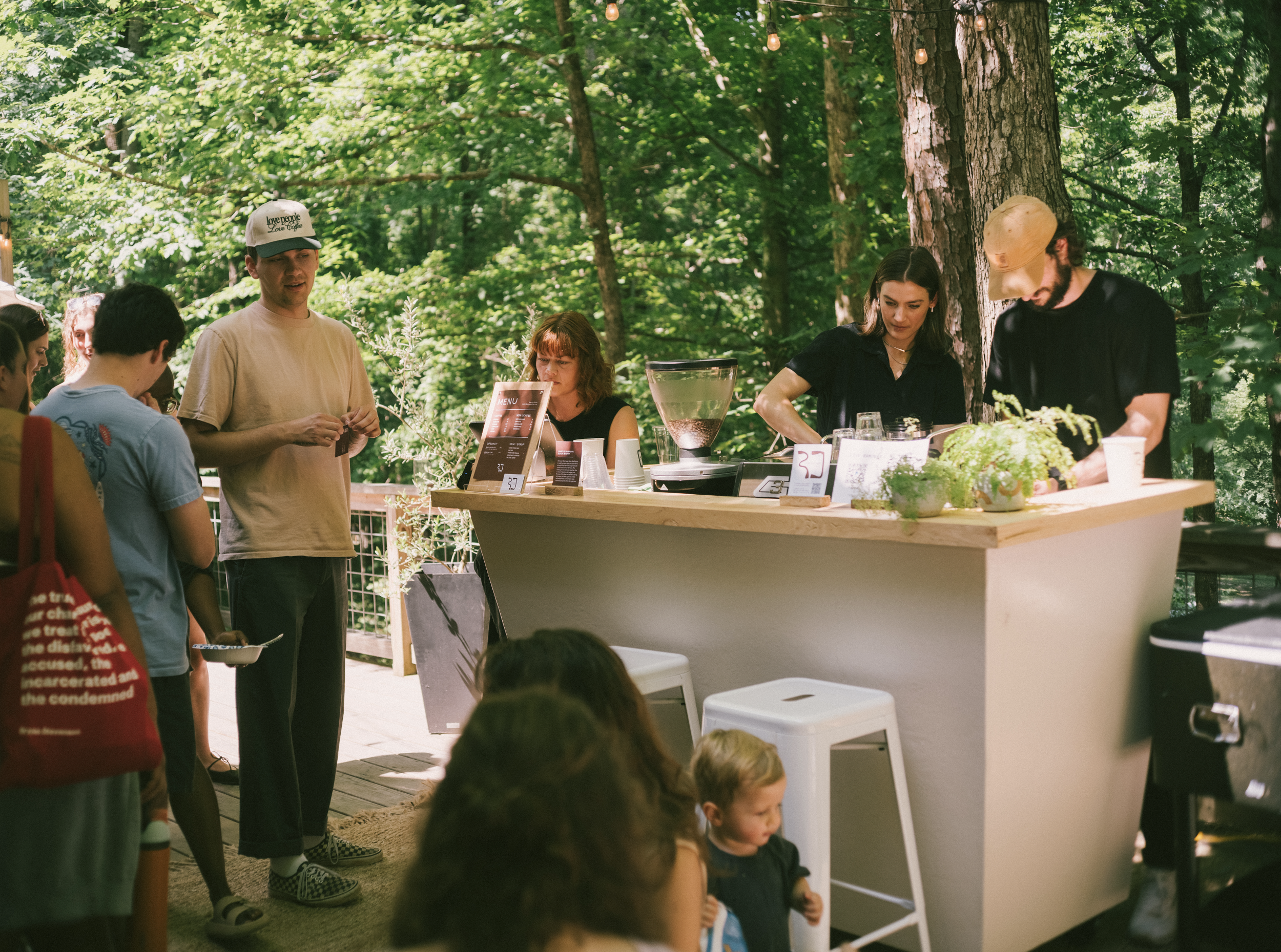 People gathering outdoors under a canopy of green trees, with a wooden bar or counter displaying various items and a menu, in a park or forest setting.