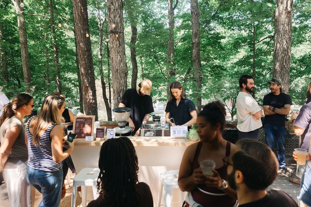 People gathered outdoors at a coffee stand in a wooded area with trees, some ordering drinks and others chatting.