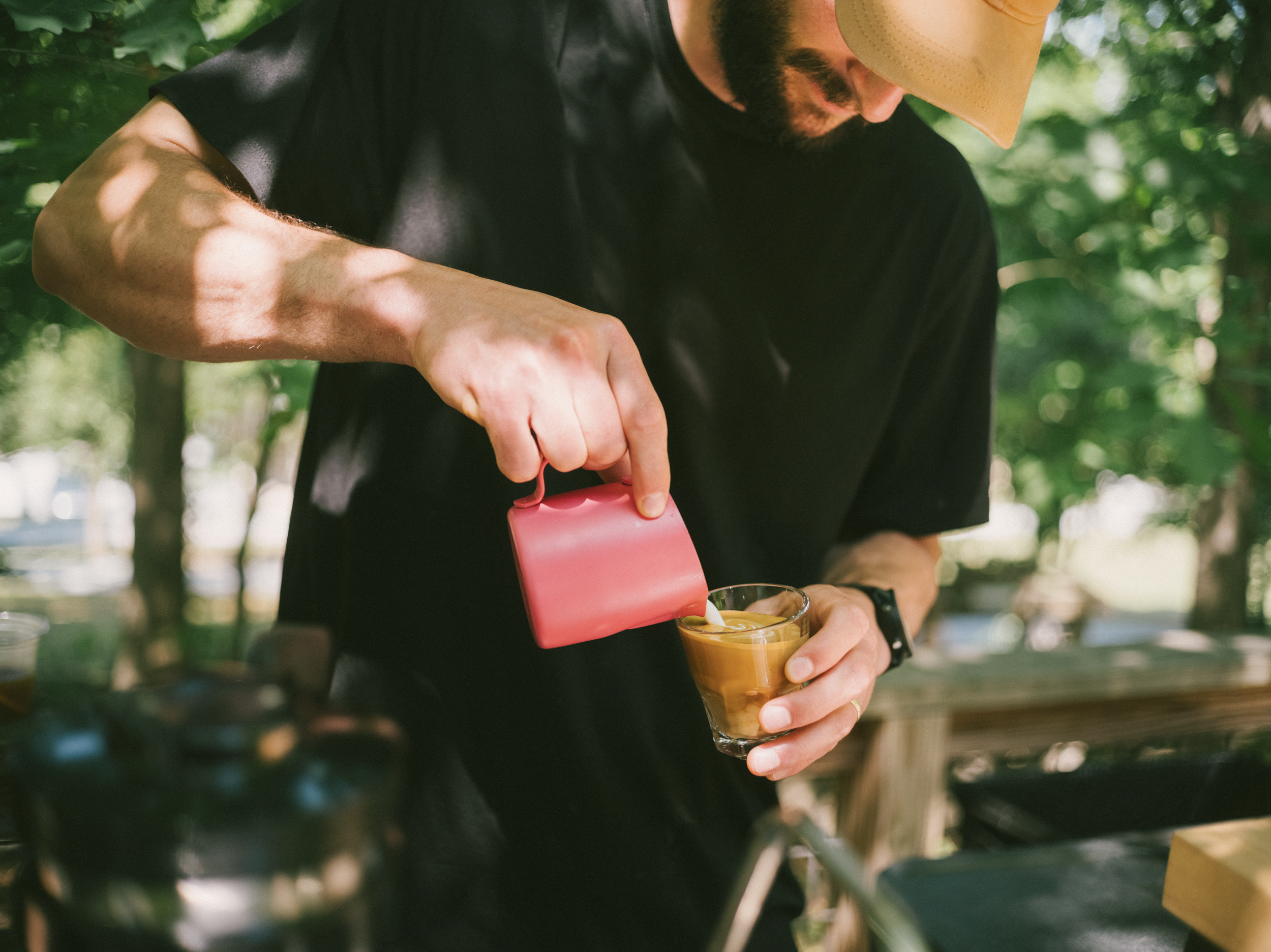 A barista wearing a black shirt pouring alternative milk from a pink milk pitcher into a clear glass with a drink, outdoors with green trees in the background.