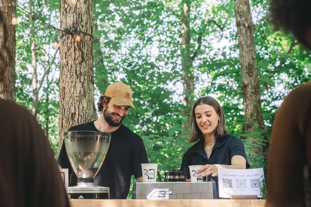 Two people are serving drinks behind a bar at an outdoor event in a wooded area, with trees and string lights overhead.