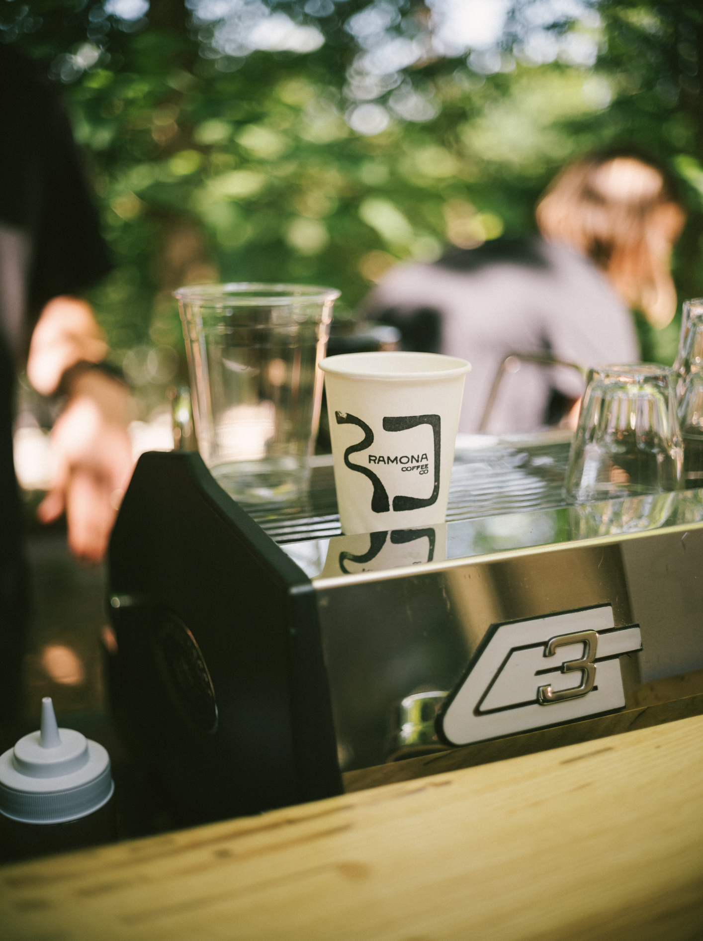 Mobile espresso cart with a La Marzocco GS3 espresso machine holding paper coffee cup labeled 'Ramona Coffee' and a clear plastic cup, with blurred background of people and trees.