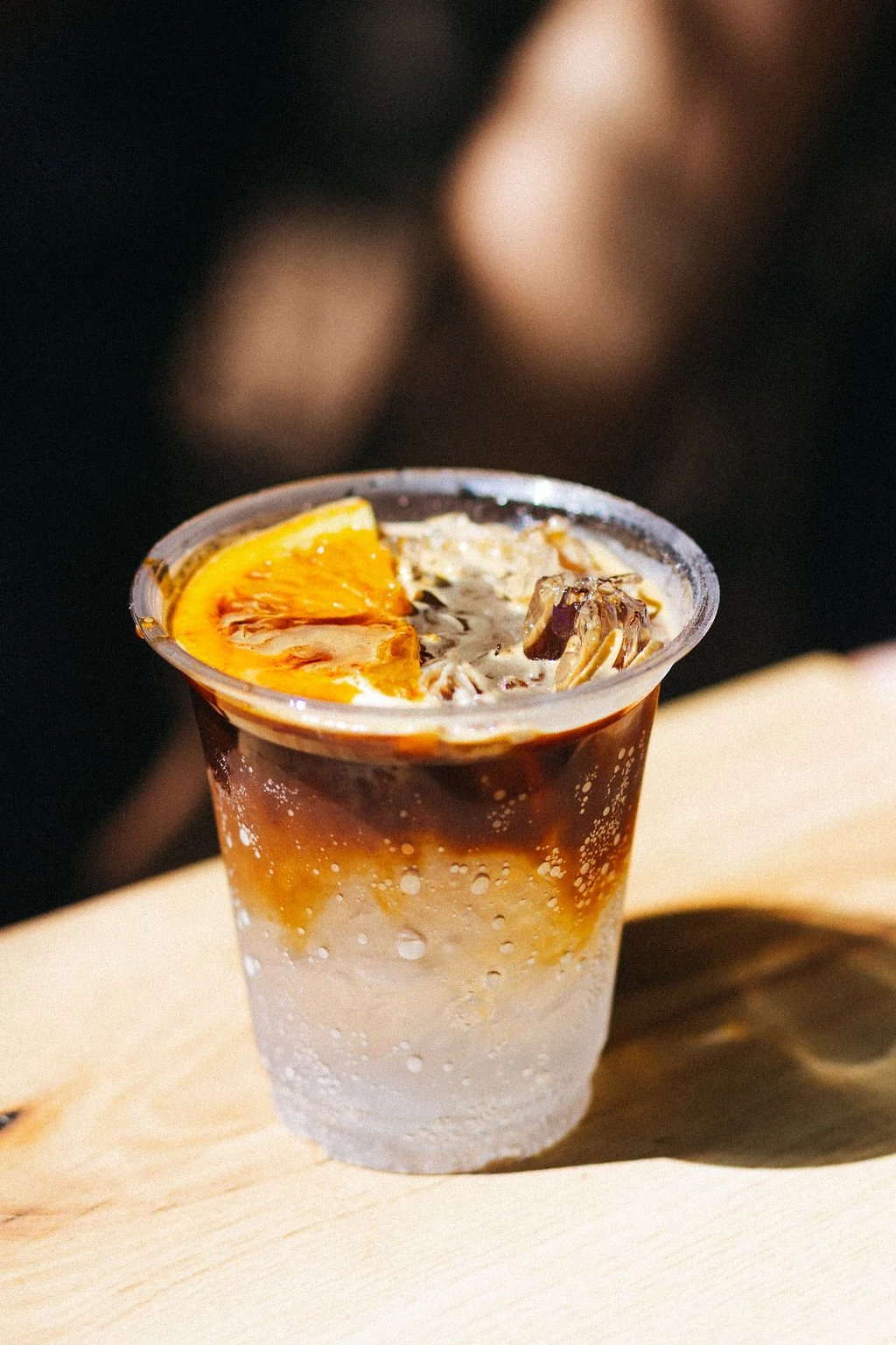 An iced espresso tonic made with espresso, tonic water, ice and a slice of orange on top, placed on a light-colored surface.