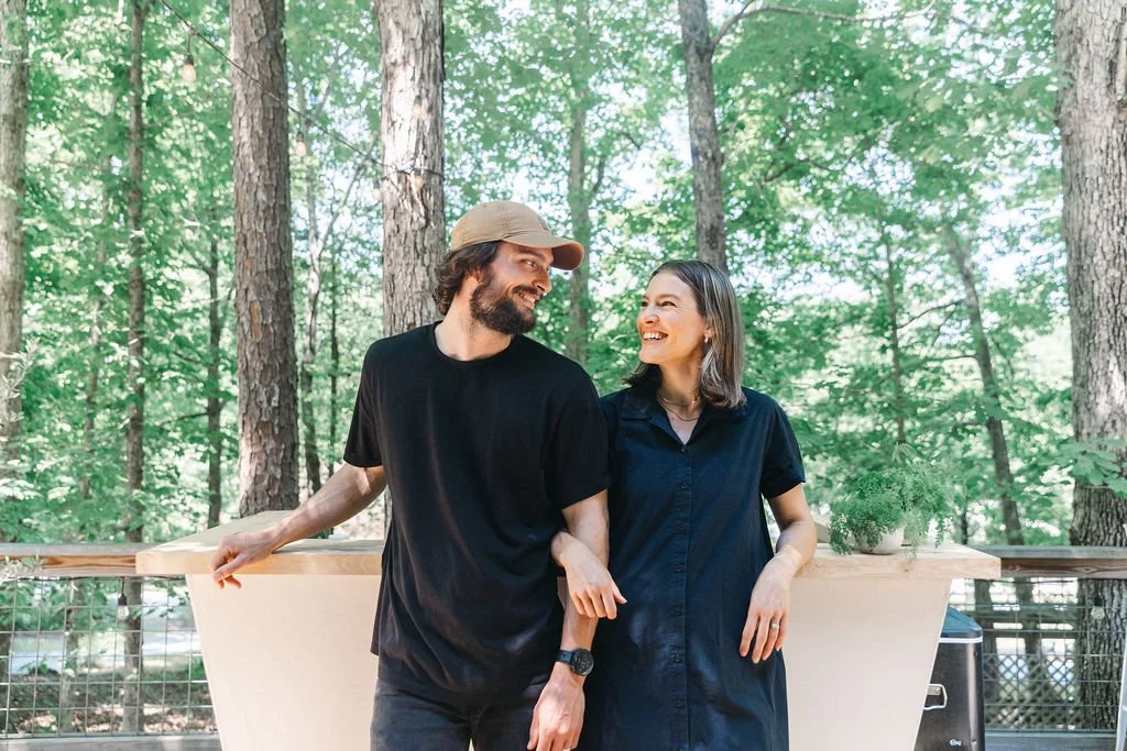 A man and woman smiling and talking while standing outdoors in a forested area with tall trees, a wooden bar, and some plants.