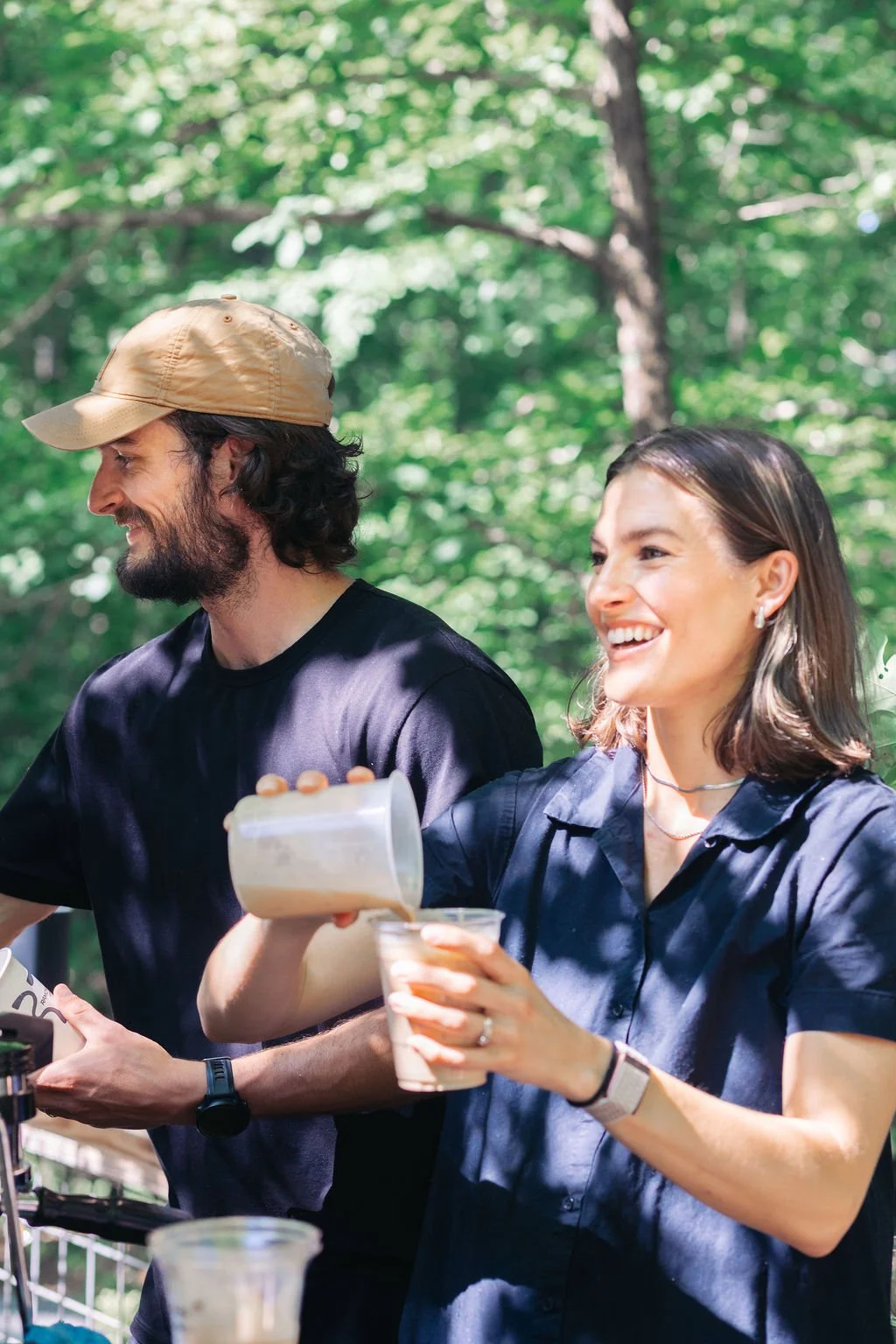A man and woman outdoors in a green wooded area, smiling and pouring drinks, enjoying a sunny day.
