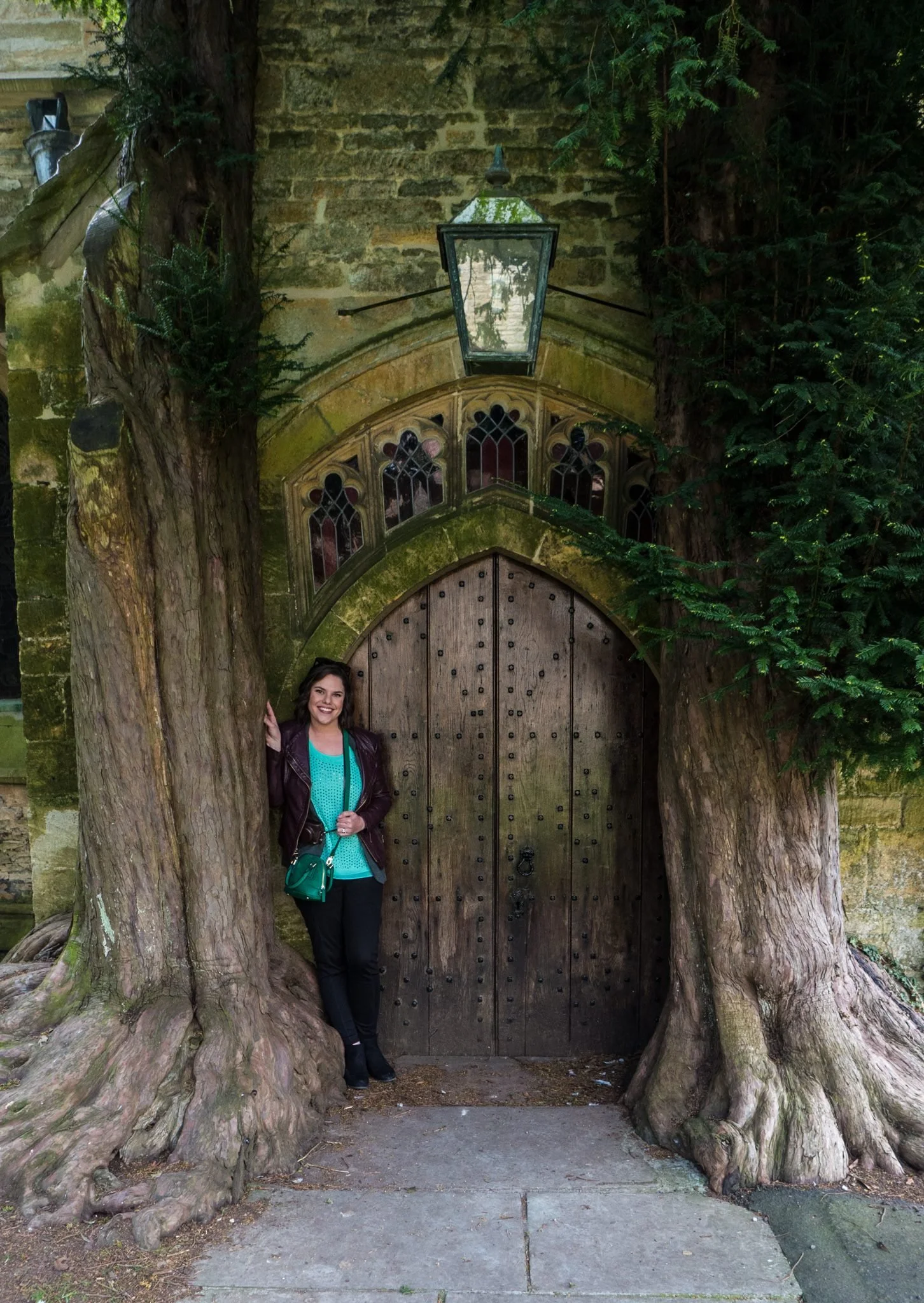 Woman with dark hair, wearing a maroon jacket over a turquoise top, standing between large tree trunks, smiling, next to a wooden door with gothic-styled windows and a lantern above it, surrounded by greenery and stone architecture.