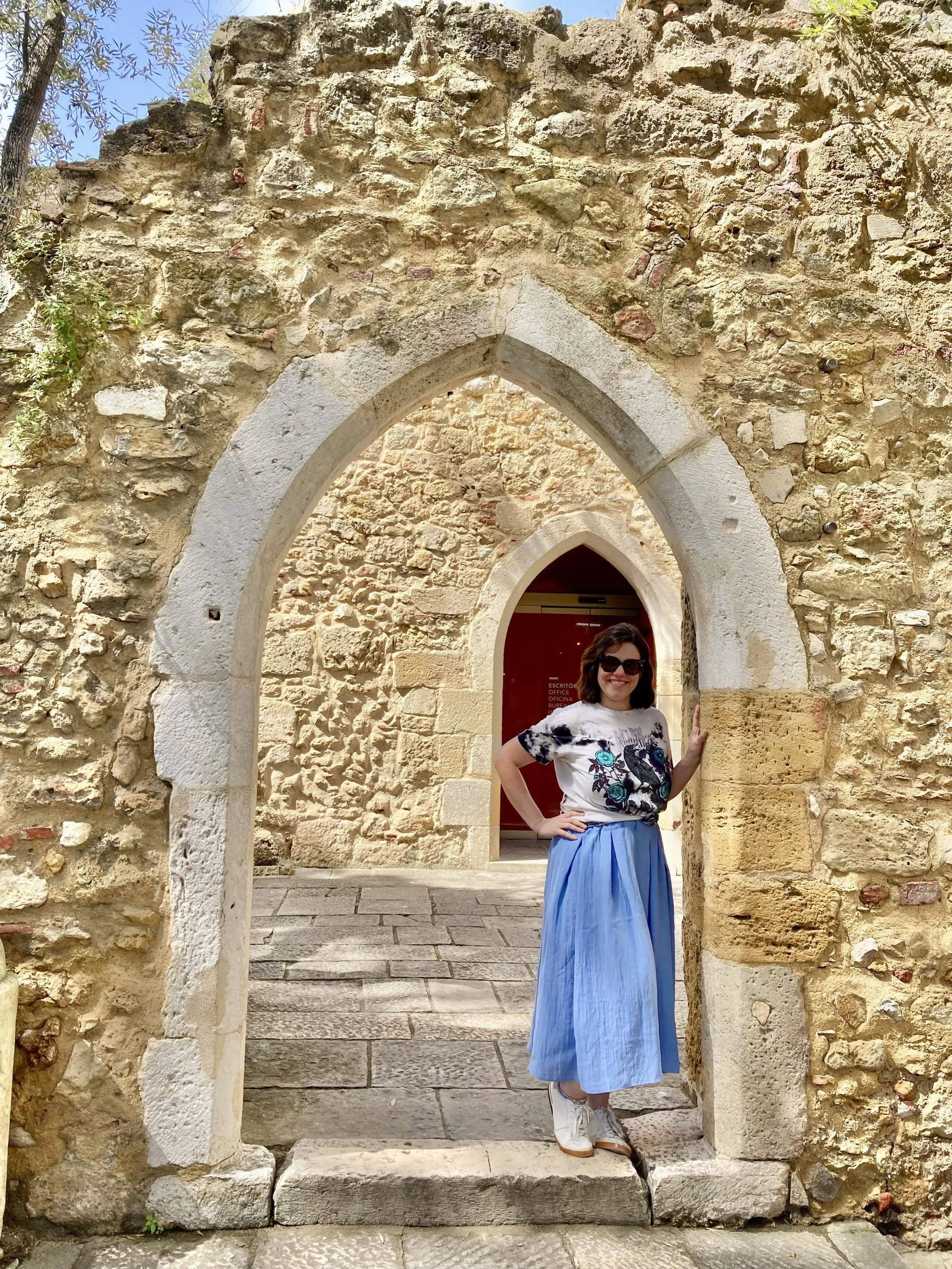 A woman standing in a doorway of an old stone archway with a weathered brick wall behind her, smiling and wearing sunglasses, a white T-shirt with a design, a long blue skirt, and white sneakers.