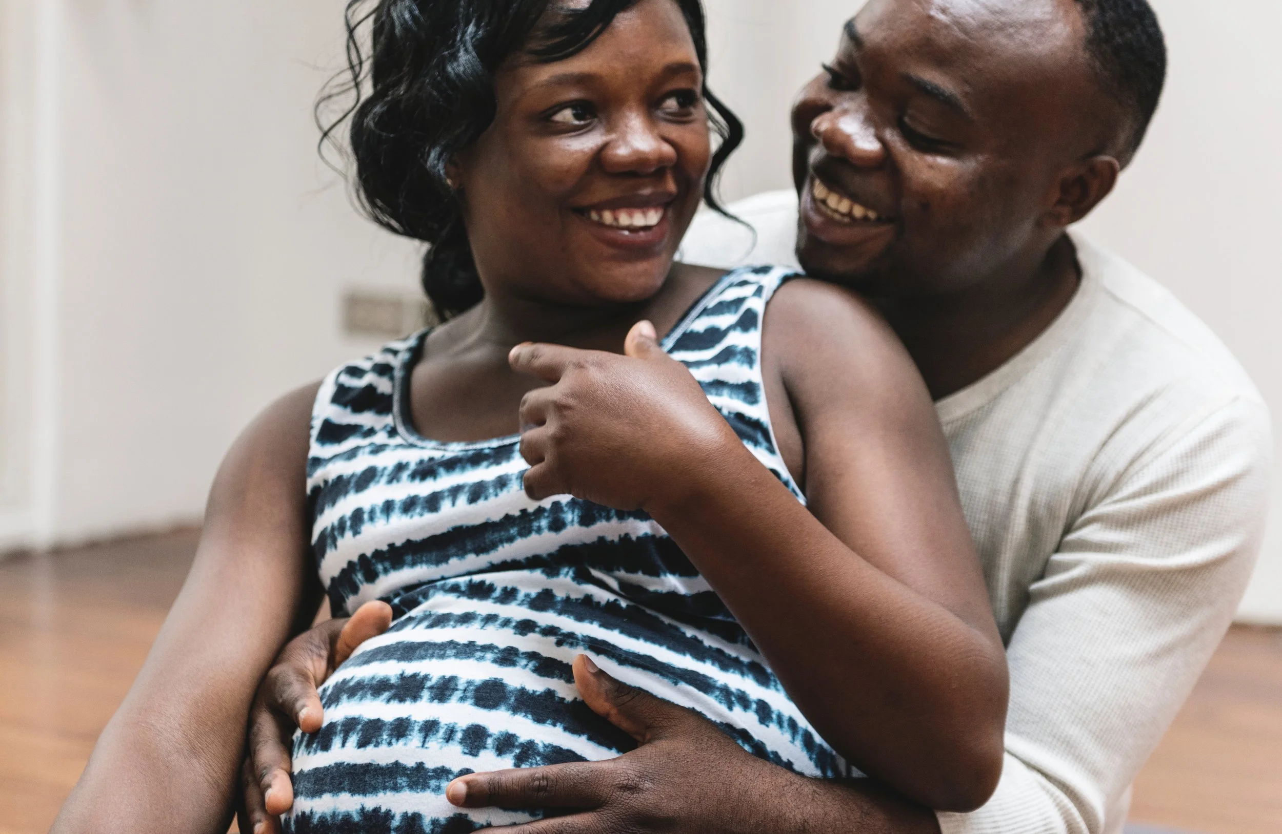 Young black pregnant couple smiling at each other whilst the man sits behind the woman with his hands on her bump