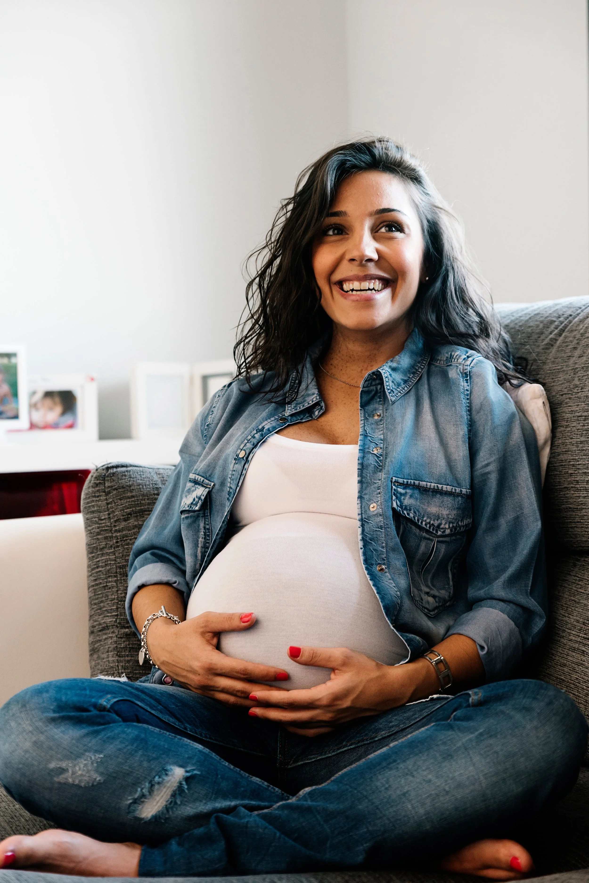 A pregnant woman with curly dark hair, wearing a denim shirt, smiling while sitting cross-legged on a sofa, holding her belly with both hands, in a cozy room with framed photos in the background.