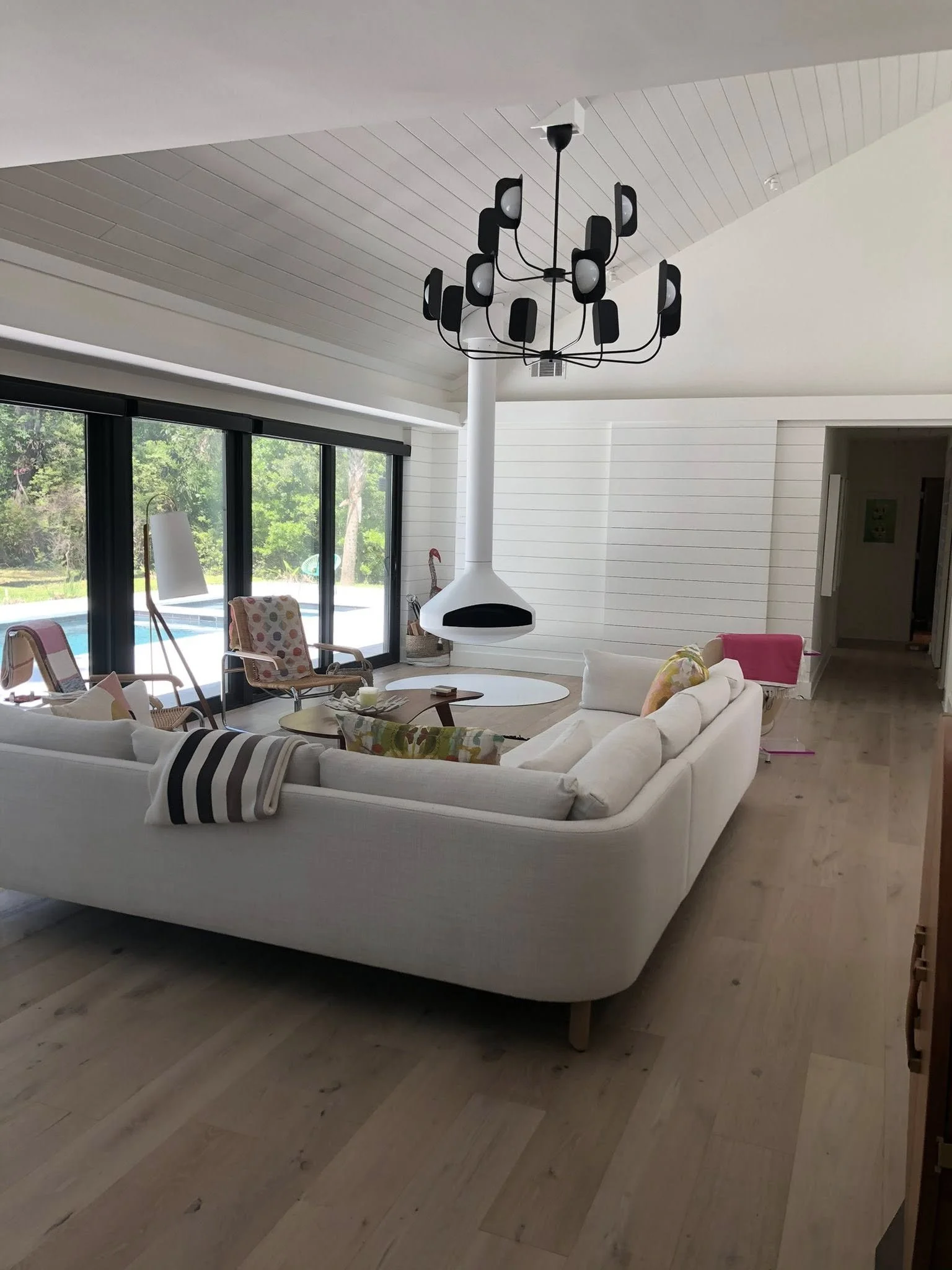A modern living room with a white sofa, armchairs with patterned cushions, a hanging black chandelier, black-framed glass sliding doors showing an outdoor pool and greenery, and white wooden panel walls.