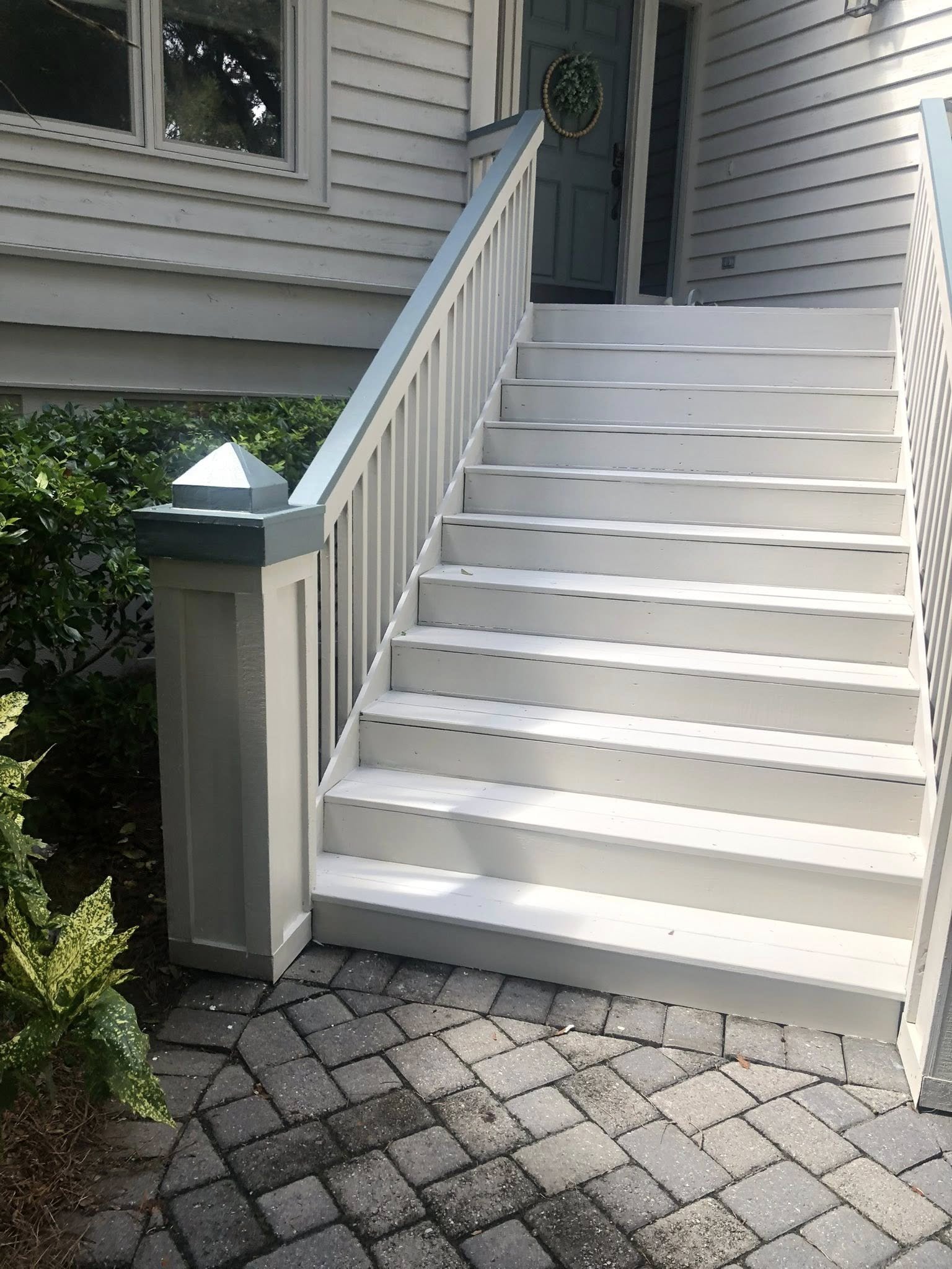 Exterior view of a white-painted wooden staircase with handrails, leading up to a front door of a house with horizontal siding. There is a window to the left and a door with a decorative wreath hanging on it. The staircase and landing are surrounded by plants and paving stones.