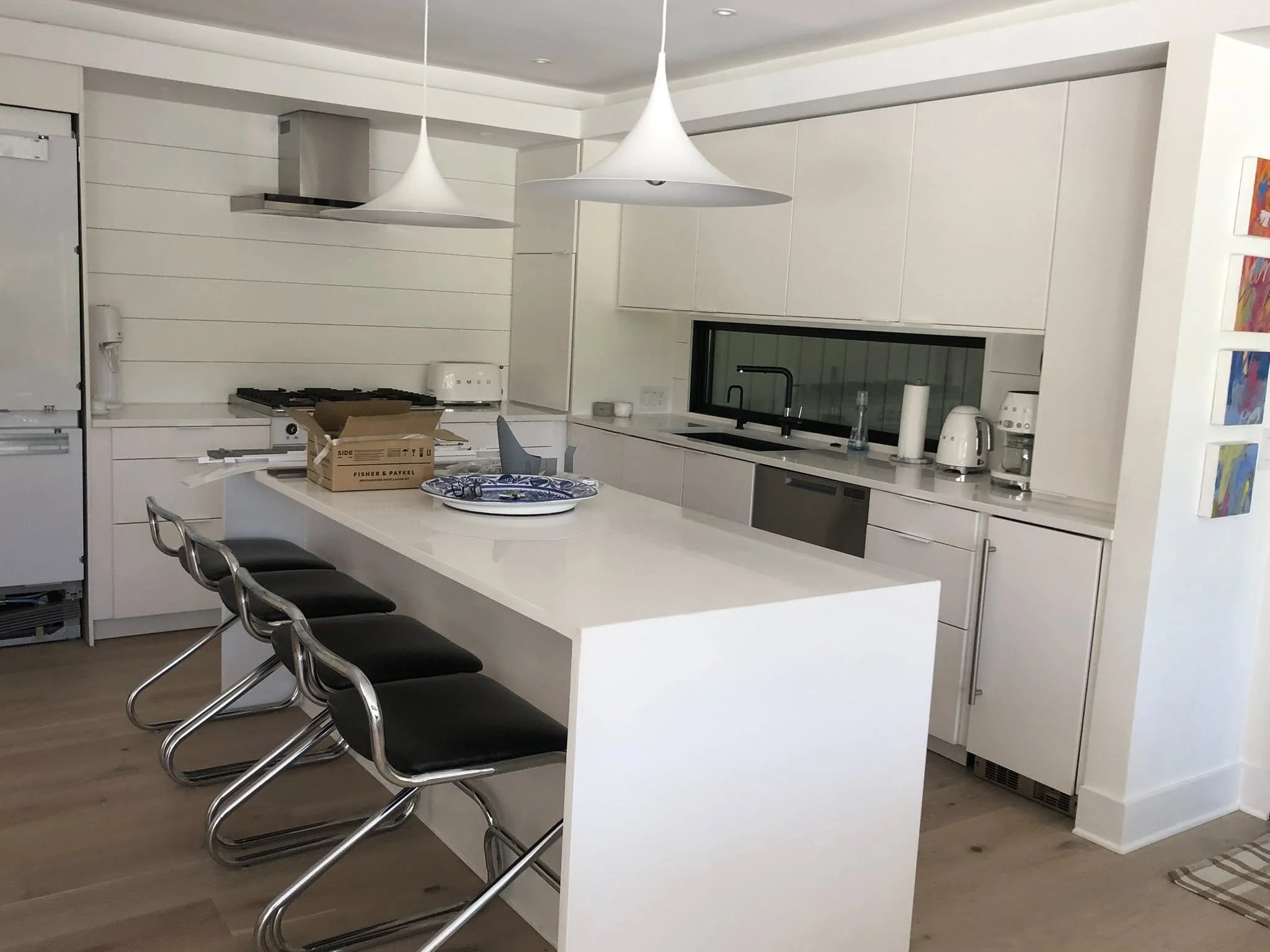 Modern white kitchen with a large countertop island, black chairs, hanging white pendant lights, and various appliances including a toaster, blender, and coffee maker.