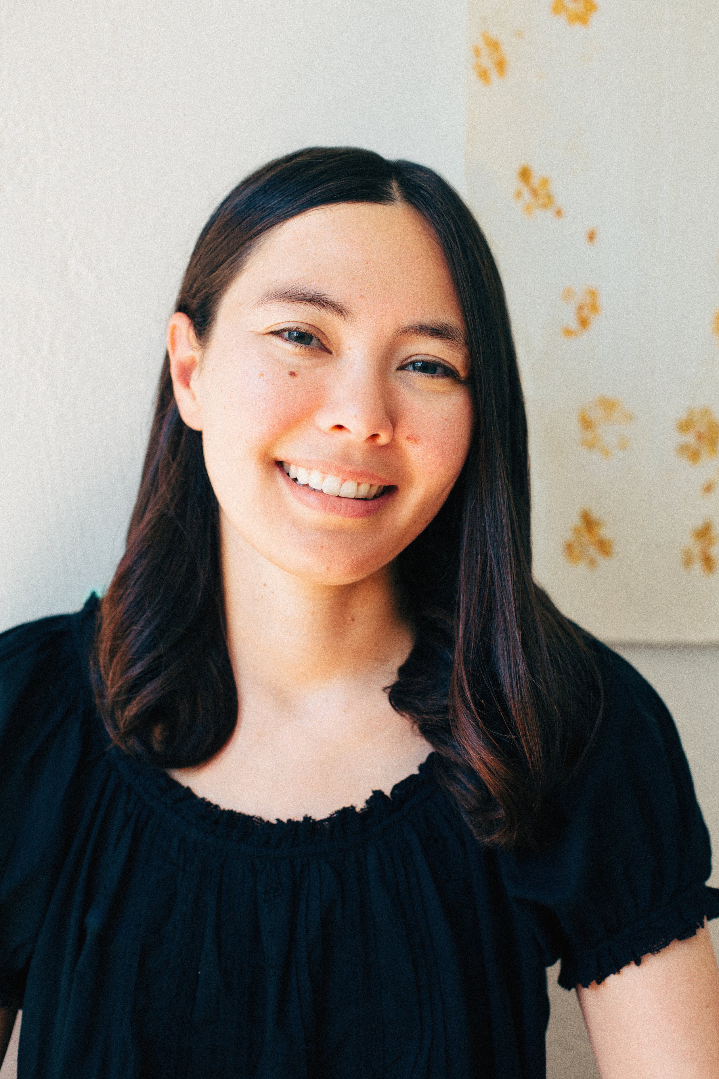 A young woman with dark hair and fair skin smiling in front of a light background with small yellow floral patterns.