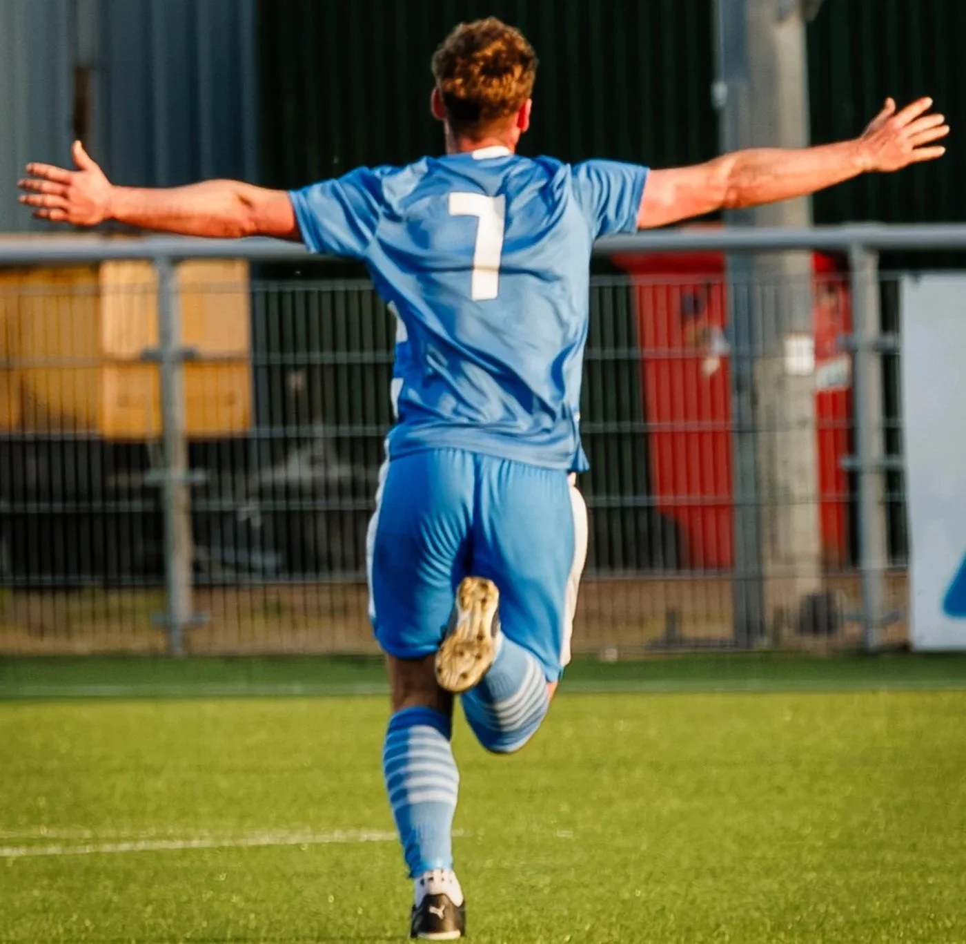 A soccer player in a blue uniform running on the field with arms outstretched, back facing the camera, wearing jersey number 7.