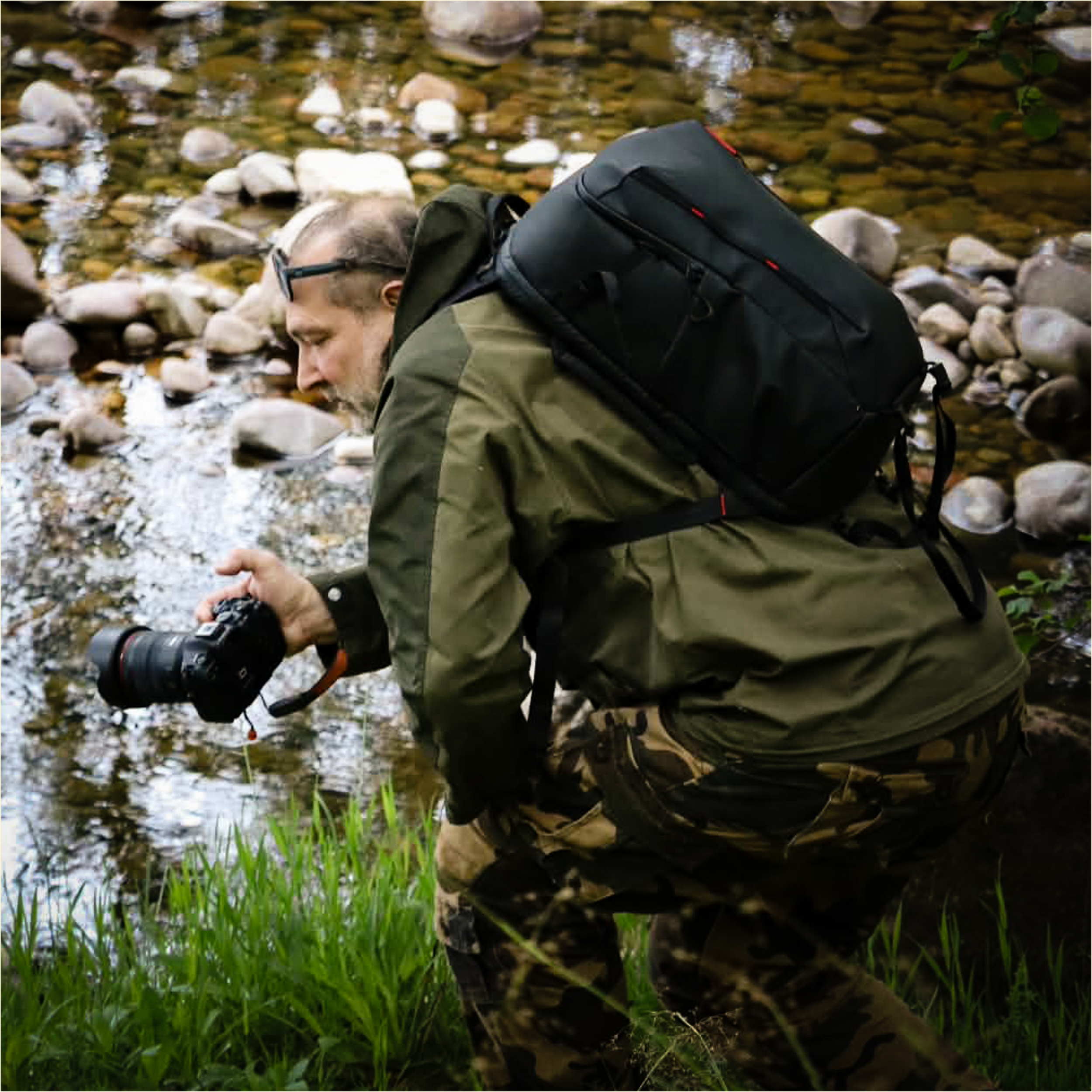 A man crouching near a rocky riverbank with water and green grass, holding a camera.