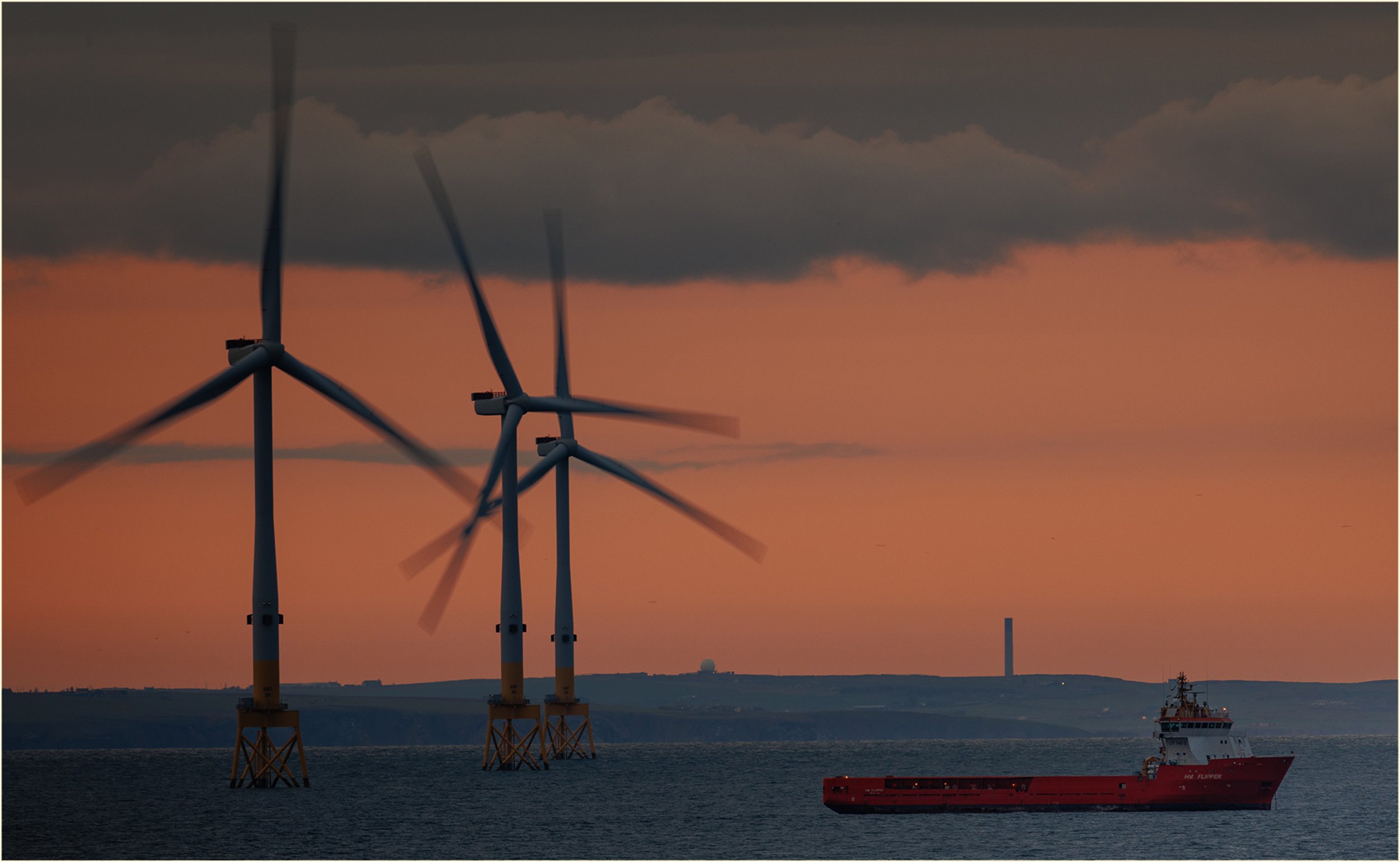 Wind turbines in the ocean during sunset with a cargo ship in the foreground and a partly cloudy sky.