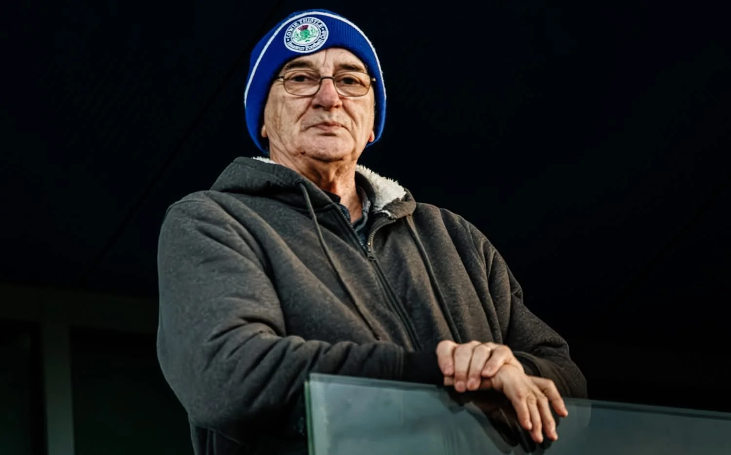 An older man wearing glasses, a blue beanie with a logo, and a dark hoodie stands with crossed arms against a dark background.
