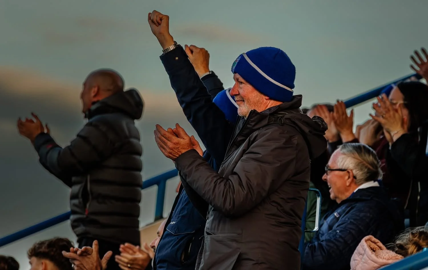 A group of people cheering at a sports event, with some standing and clapping, and one man raising his fist in celebration, outdoors during the evening or late afternoon.