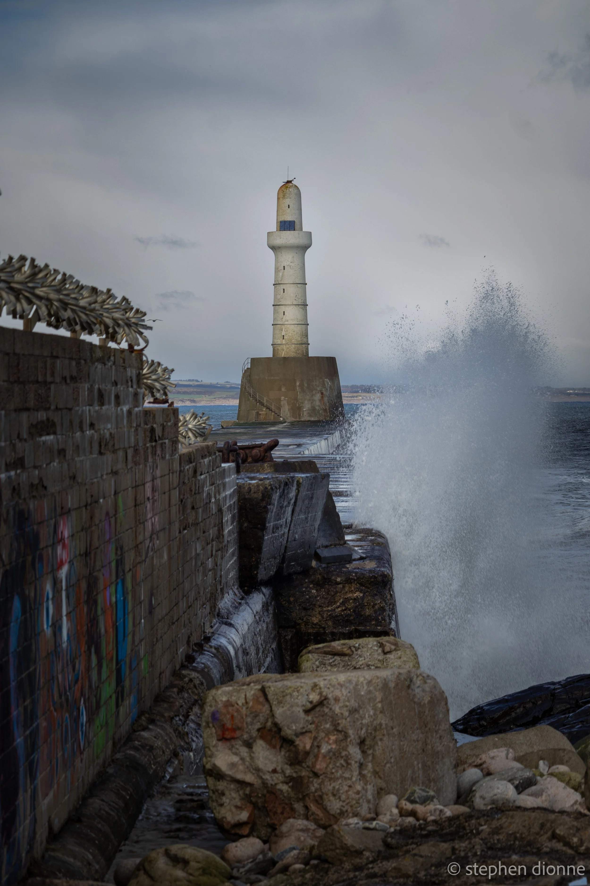 A lighthouse situated on a concrete base at the edge of the water, with waves crashing against it, creating spray. The sky is overcast with dark clouds, and there's a graffiti-covered brick wall on the left side of the image, with rocks and pebbles in the foreground.
