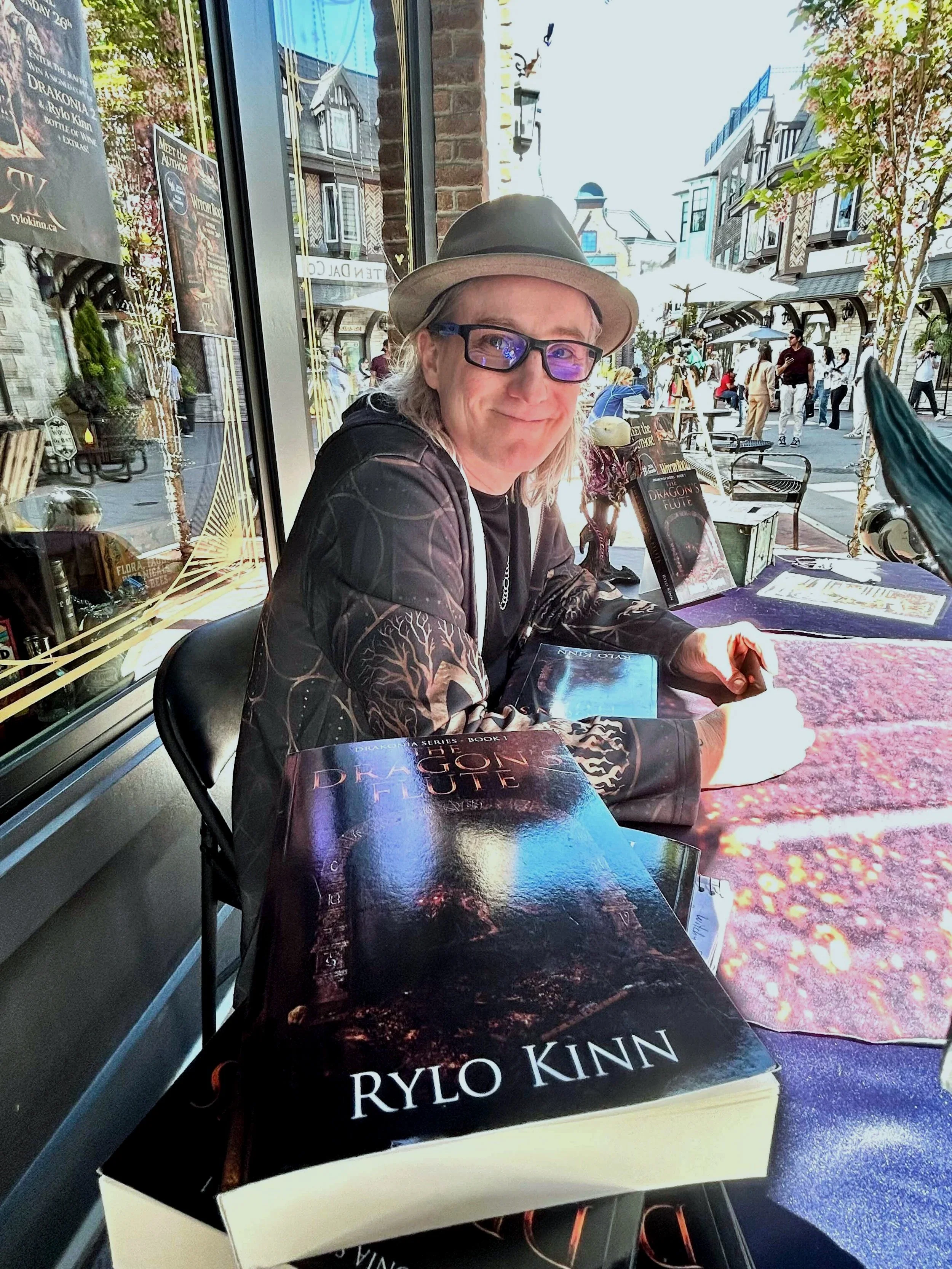 The author Rylo Kinn, sitting outside in the sun, at a table. Pictured next to Book 1 of the Drakonia Series, The Dragon's Flute.