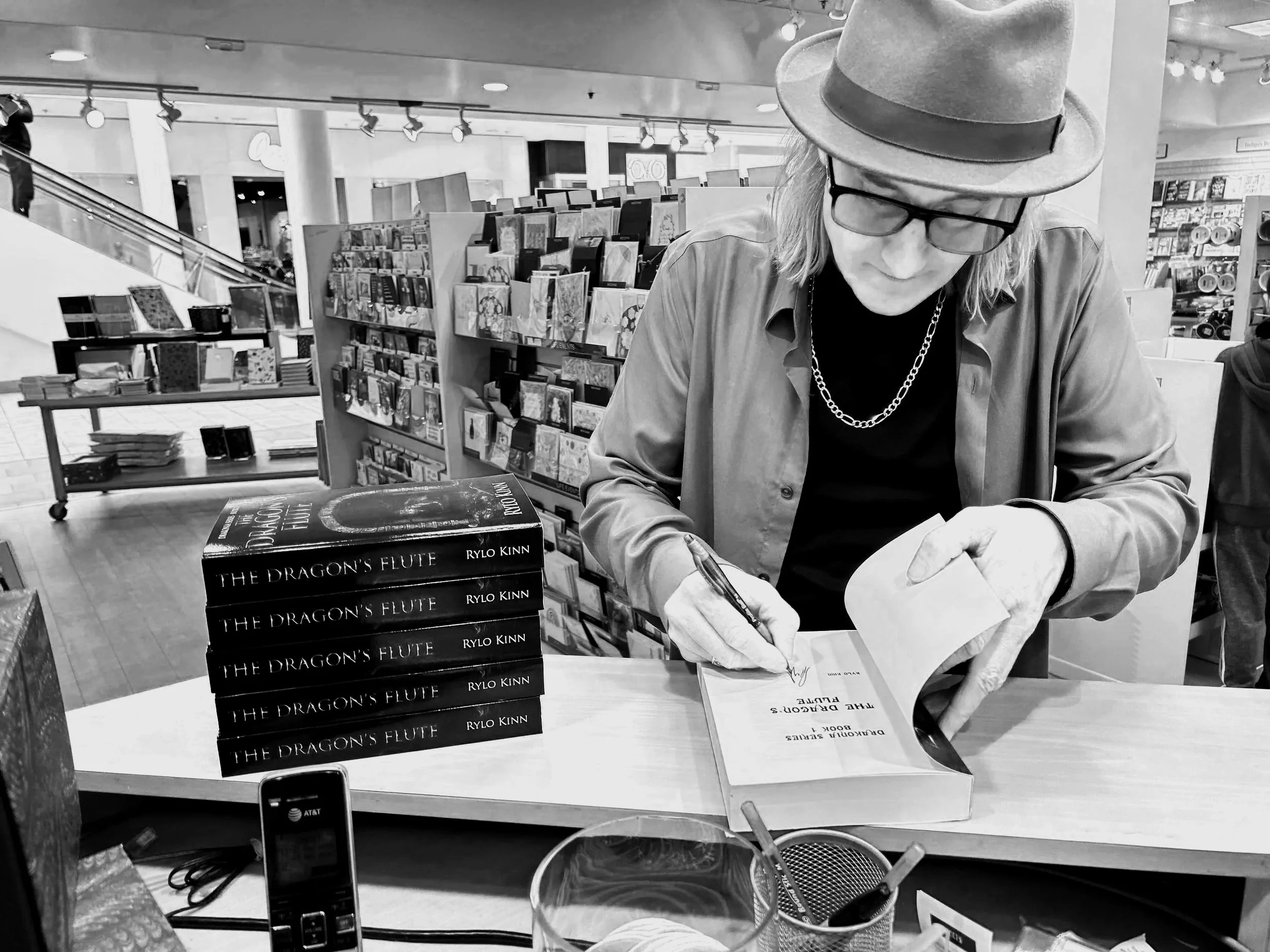 The author, Rylo Kinn, wearing glasses, a hat, and a chain necklace signing a book at a bookstore event. Multiple copies of the book titled "The Dragon's Flute" by Rylo Kinn are stacked on the table in front of them.