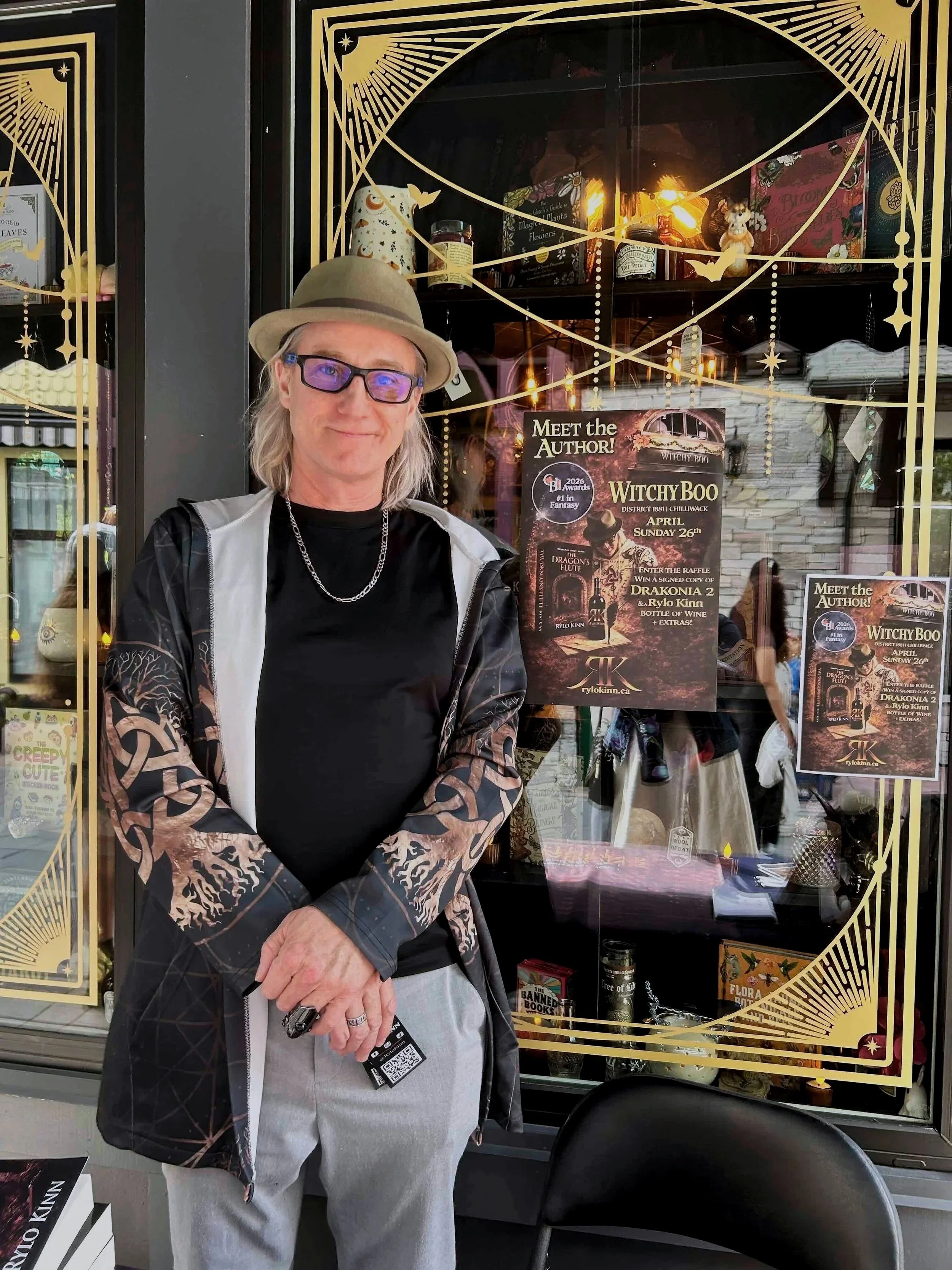A man with long gray hair wearing a beige hat, glasses, a black t-shirt, and a patterned jacket standing in front of a bookshop window. The window features promotional posters for a book signing event with the author of 'Witchy Boo,' with shelves displaying books and decorative items behind him.