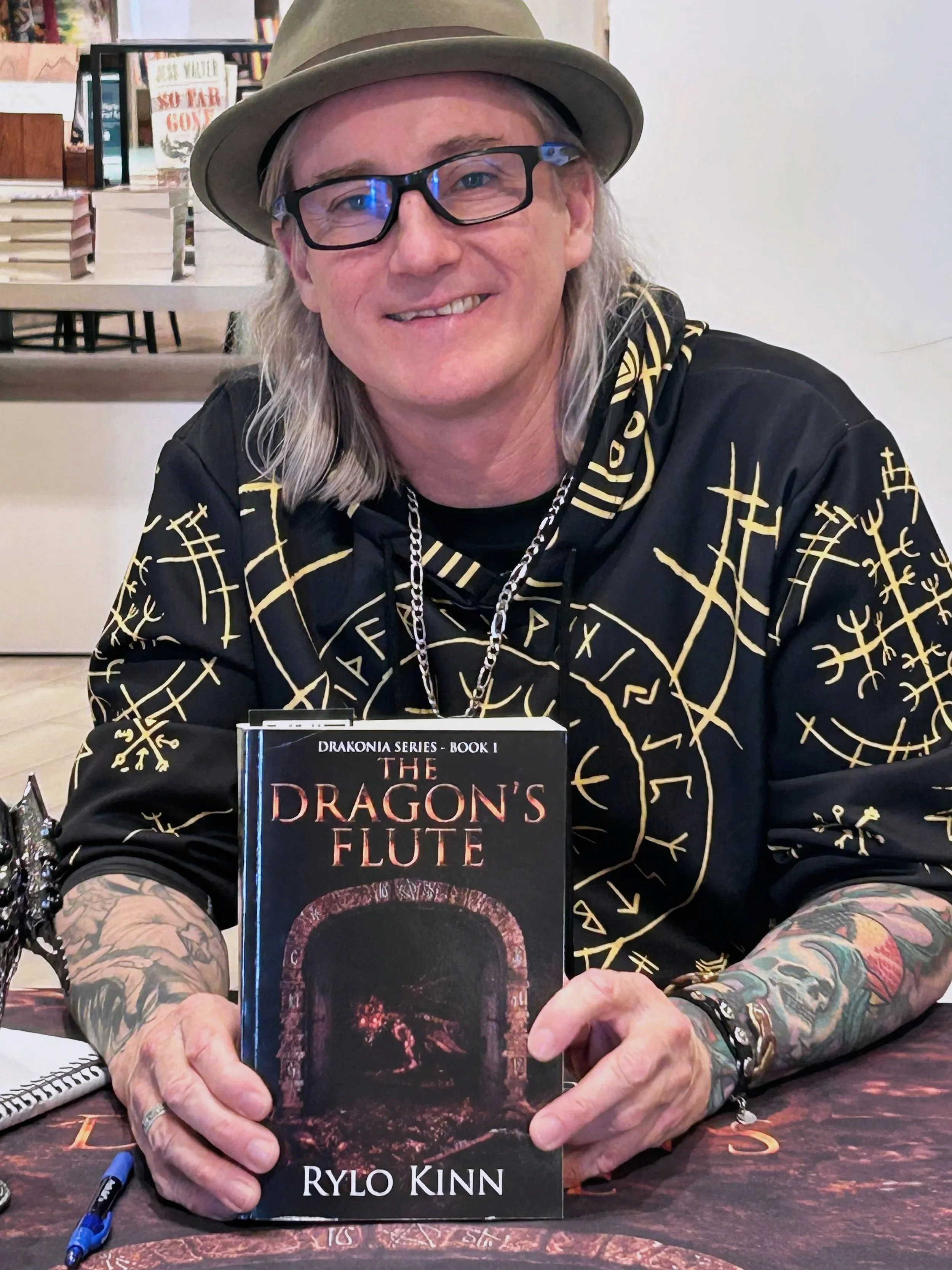 Author, Rylo Kin with long gray hair, glasses, and tattoos on his arms, wearing a fedora hat and black hoodie with yellow symbols, is sitting at a table holding a book titled "The Dragon's Flute" by Rylo Kinn. He is smiling and looking at the camera.