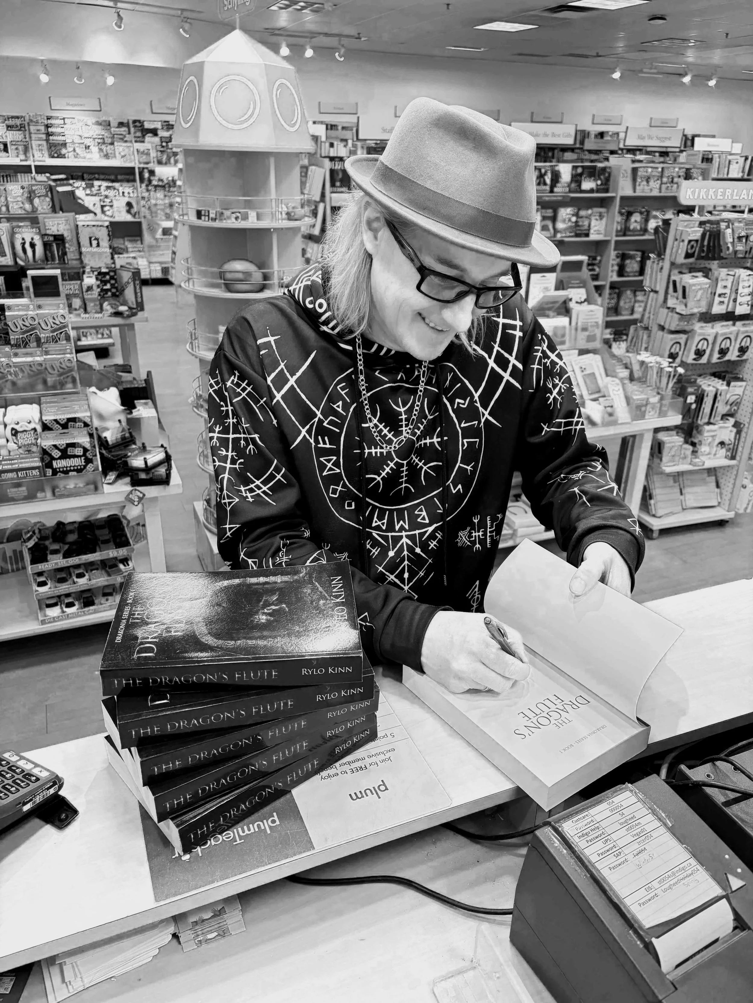 Author, Rylo Kinn, with glasses and a hat signing a book at a bookstore counter. Several copies of the book titled 'The Dragon's Flute' by Rylo Kinn are stacked on the counter. Bookshelves filled with various books are in the background.