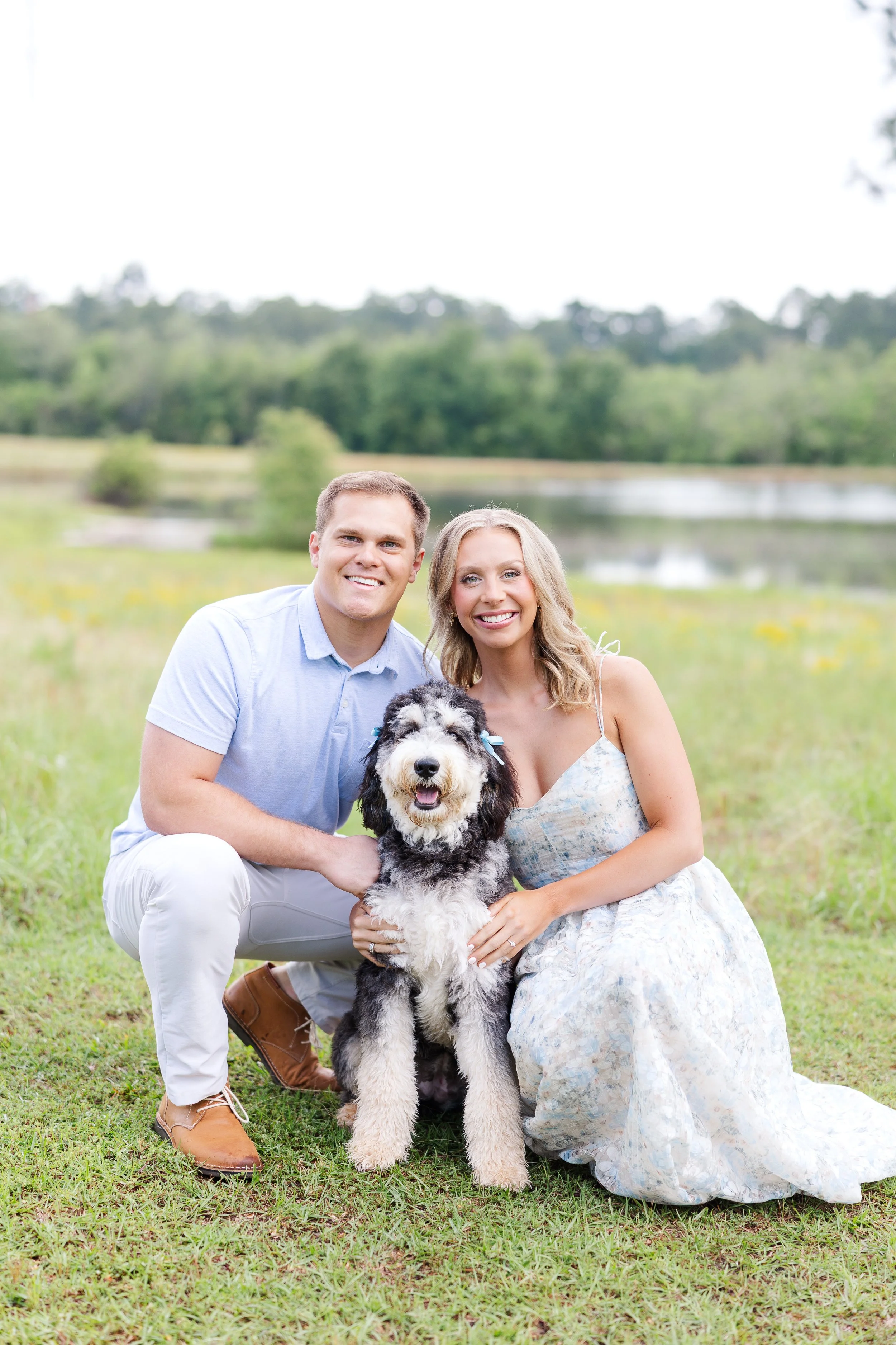 A happy couple sits outdoors in a field with green grass and trees, holding a large black, white, and gray fluffy dog. The man wears a light blue shirt and white pants, while the woman wears a flowy summer dress. The background includes a lake and distant trees.
