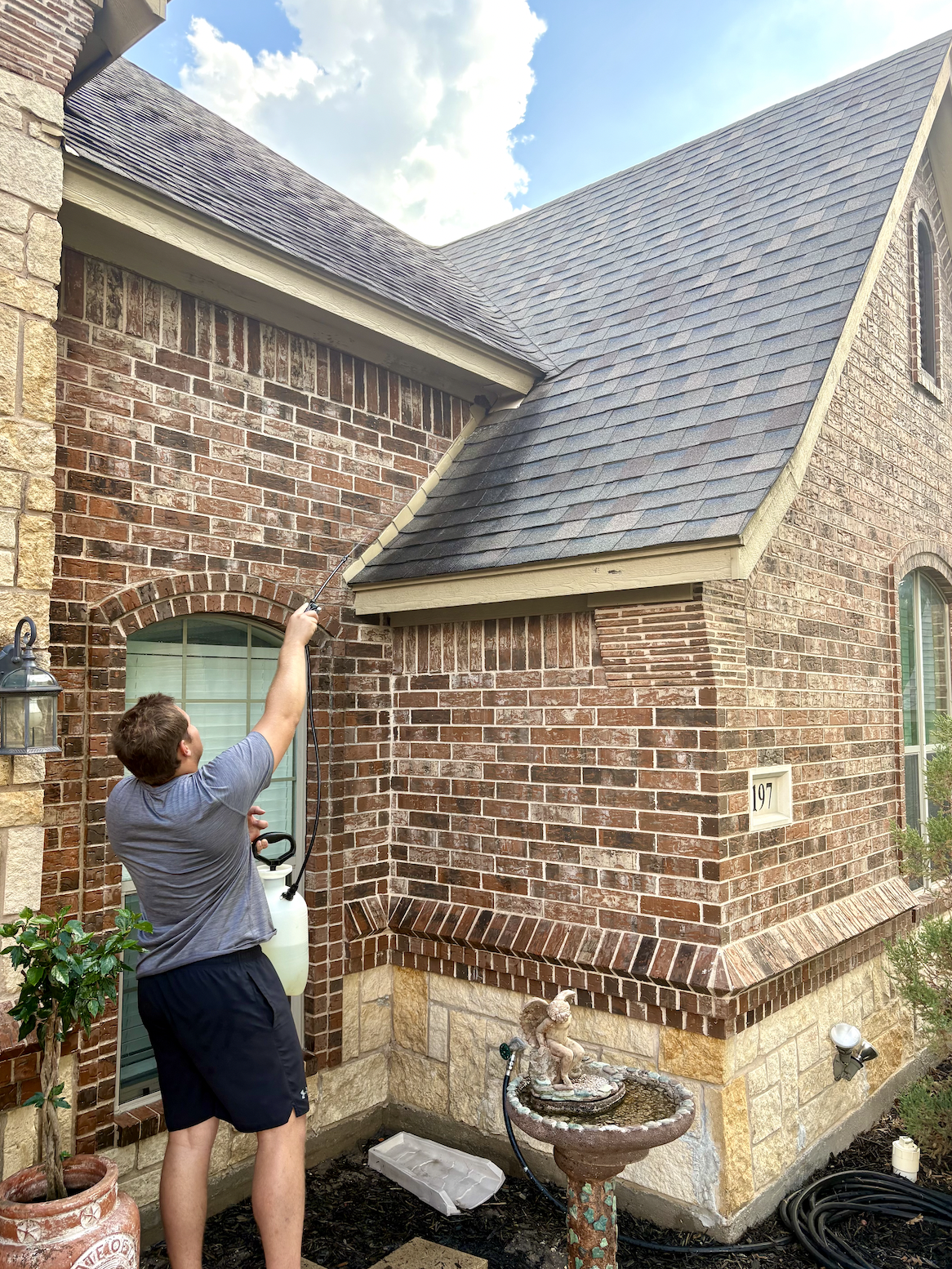 A young man is spraying the exterior brick wall of a house with a hose, aiming at the roof.