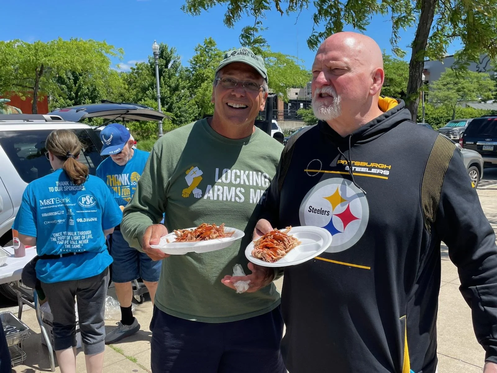Craig Wolfley and Leo Wisniewski smiling and holding plates of food at the Light of Life Rescue Mission Walk for the Homeless, with others in the background, two women wearing blue shirts with sponsor logos.