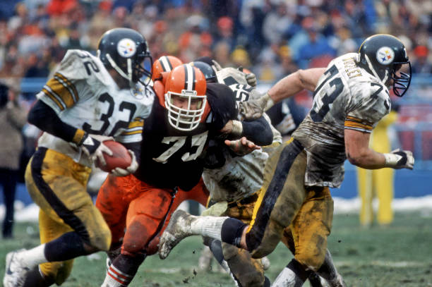 Craig Wolfley and Franco Harris in action during a game, with players from the Pittsburgh Steelers in white and black jerseys and an opposing player in a black jersey with orange pants, on a football field.