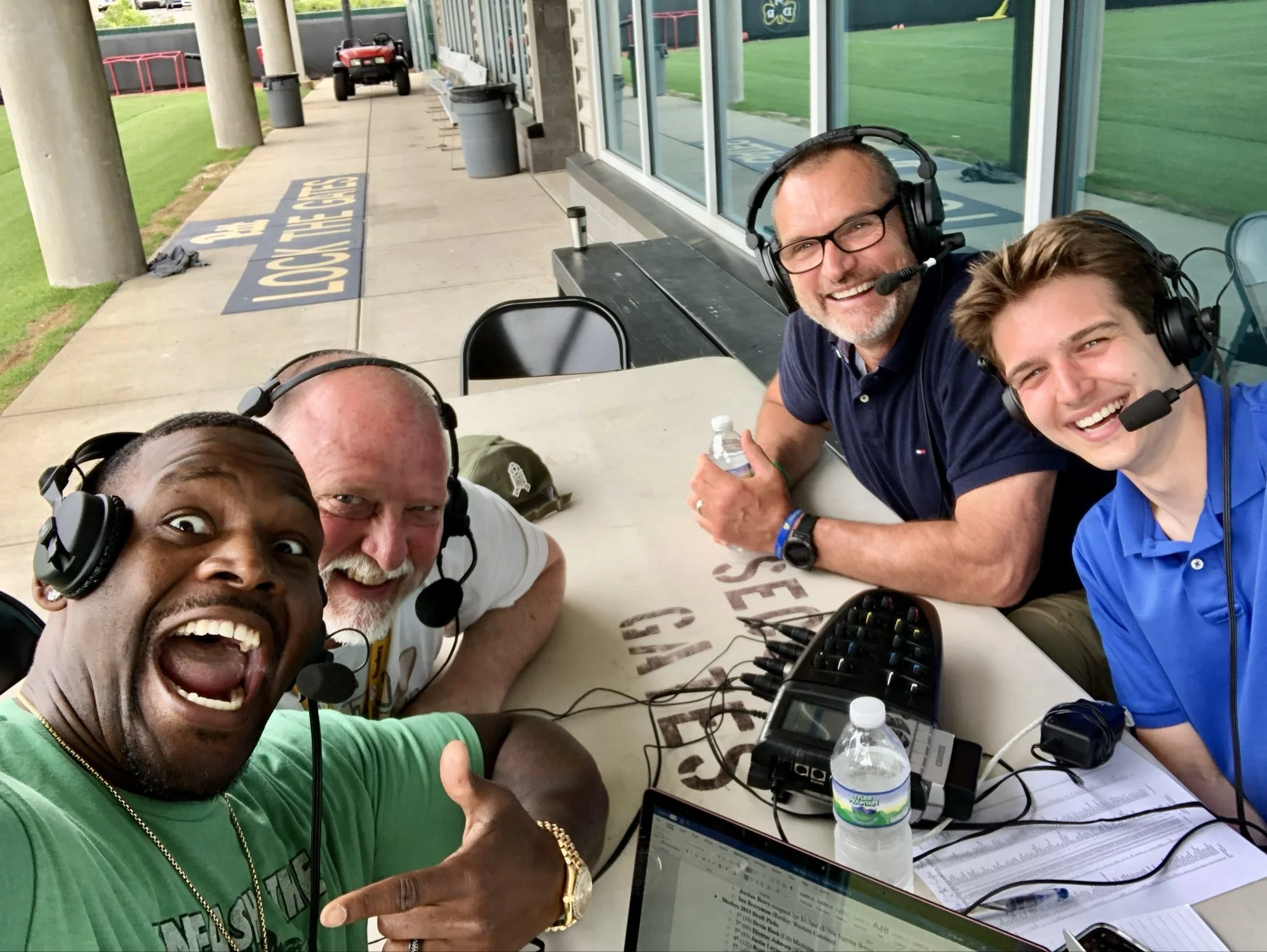 Craig Wolfley, Tunch Ilkin, Wes Uhler and Arthur Moats wearing headsets with microphones, smiling, sitting at a table outdoors, with electronic equipment, laptops, and water bottles, possibly during a sports event or broadcast.