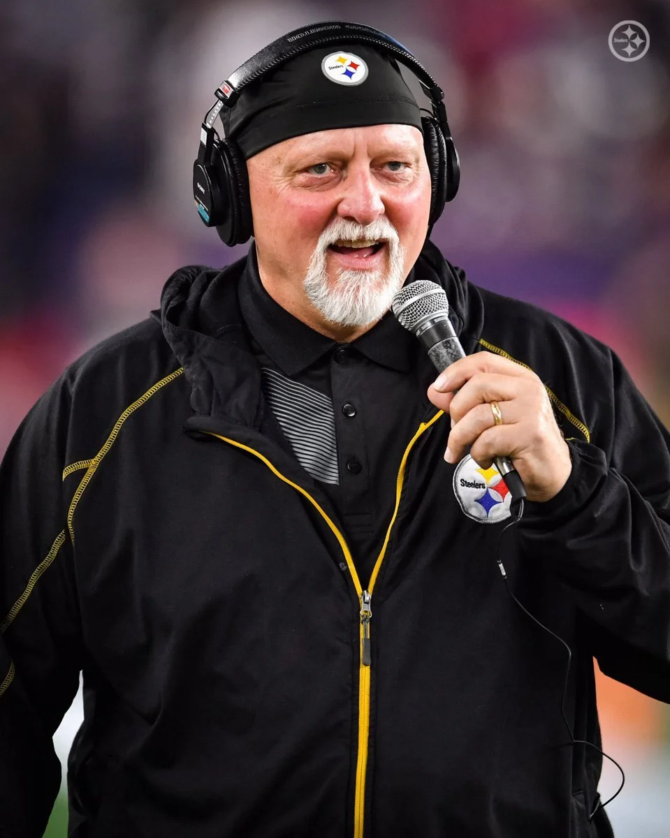 Craig Wolfley wearing a black Pittsburgh Steelers jacket and headband, holding a microphone and speaking.