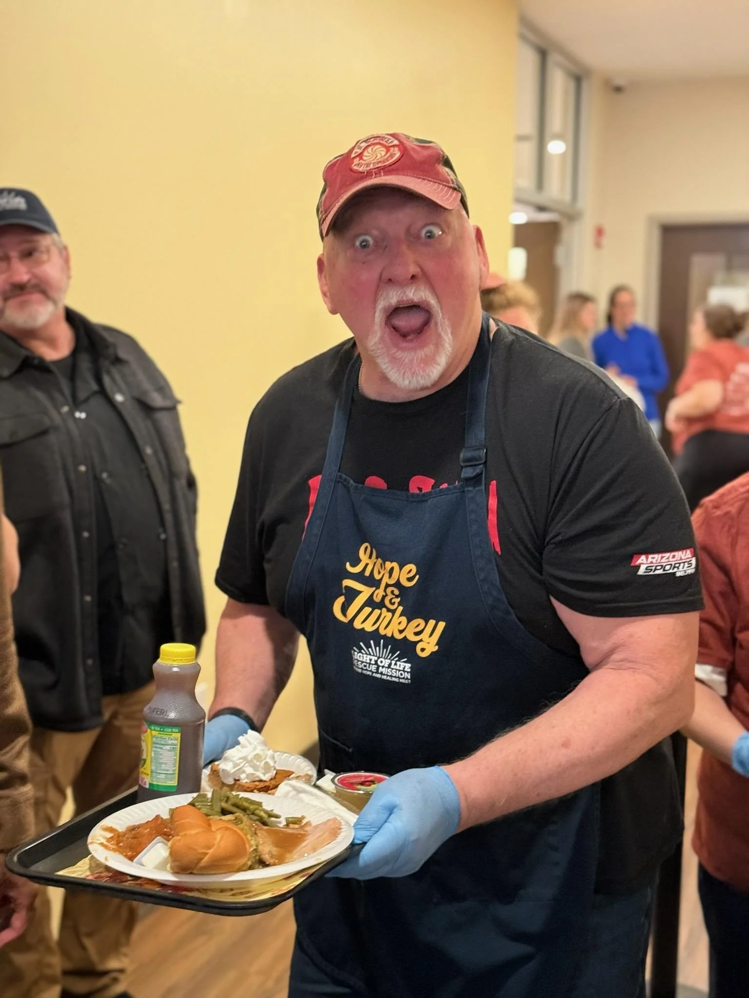 Craig Wolfley wearing a red cap and a black shirt with the phrase 'Hope & Turkey' on it is serving food on a tray on Thanksgiving Day at the Light of Life Rescue Mission. He is wearing blue gloves and has an open mouth expression.