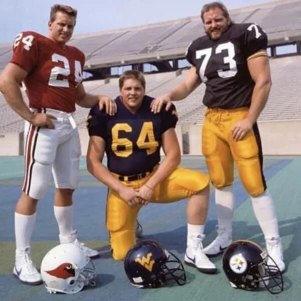 Three brothers, Craig, Ron and Dale Wolfley, in football uniforms posing on a football field during daytime, with helmets in front of them. One is kneeling, and two are standing beside him.