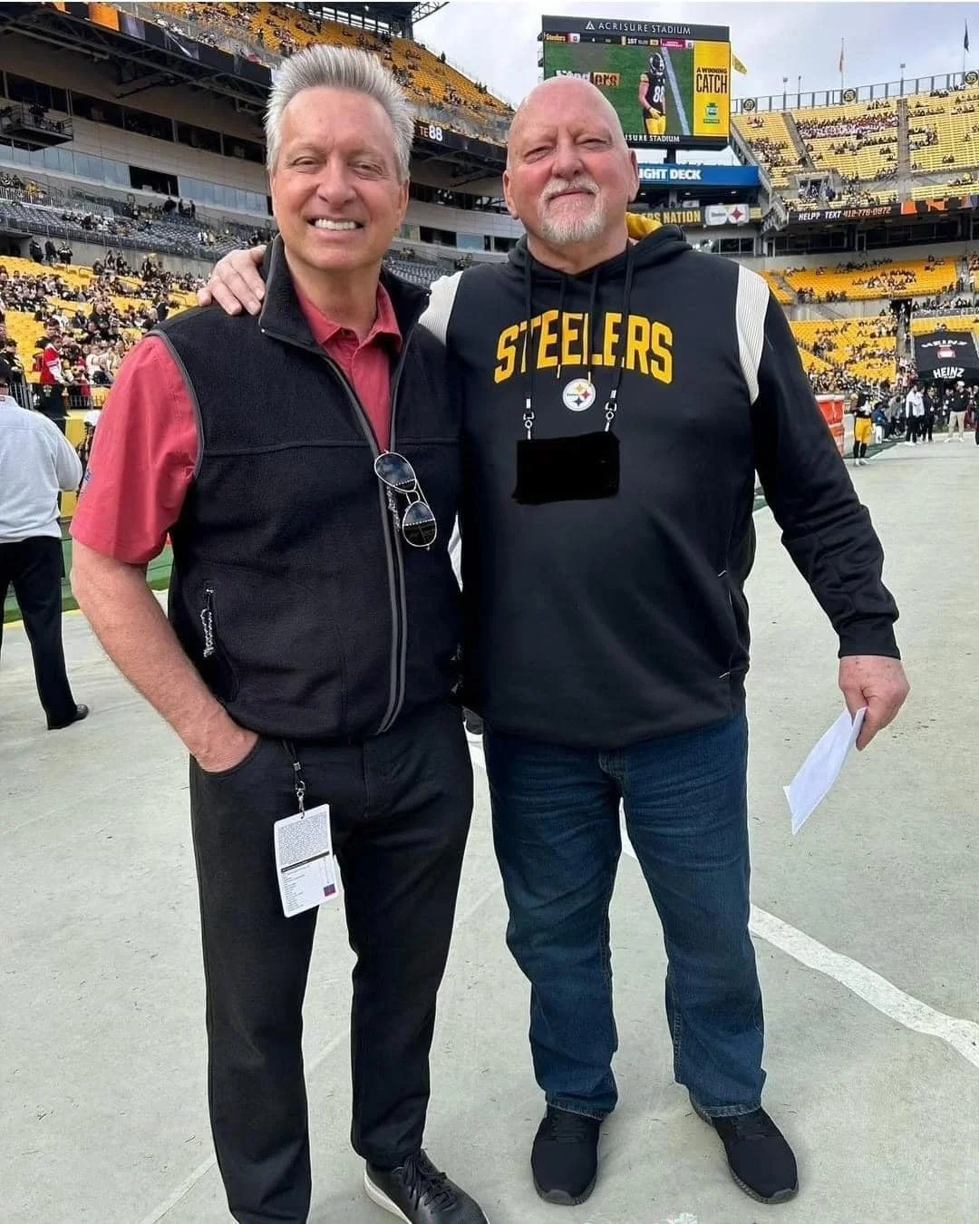Craig and Ron Wolfley standing on a football field at Heinz Field, with a Steelers hoodie and a vest, smiling and posing for a photo, with a crowd and stadium in the background.