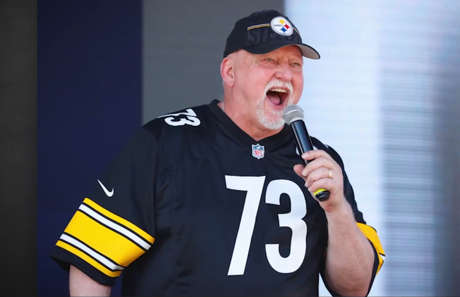 Craig Wolfley wearing a black Pittsburgh Steelers football jersey with number 73 and a Steelers cap, holding a microphone and speaking.