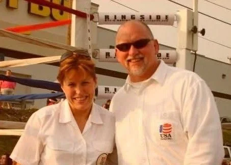 Craig and Faith Wolfley wearing white shirts with USA-themed logos, smiling outdoors with a boxing ring in the background.