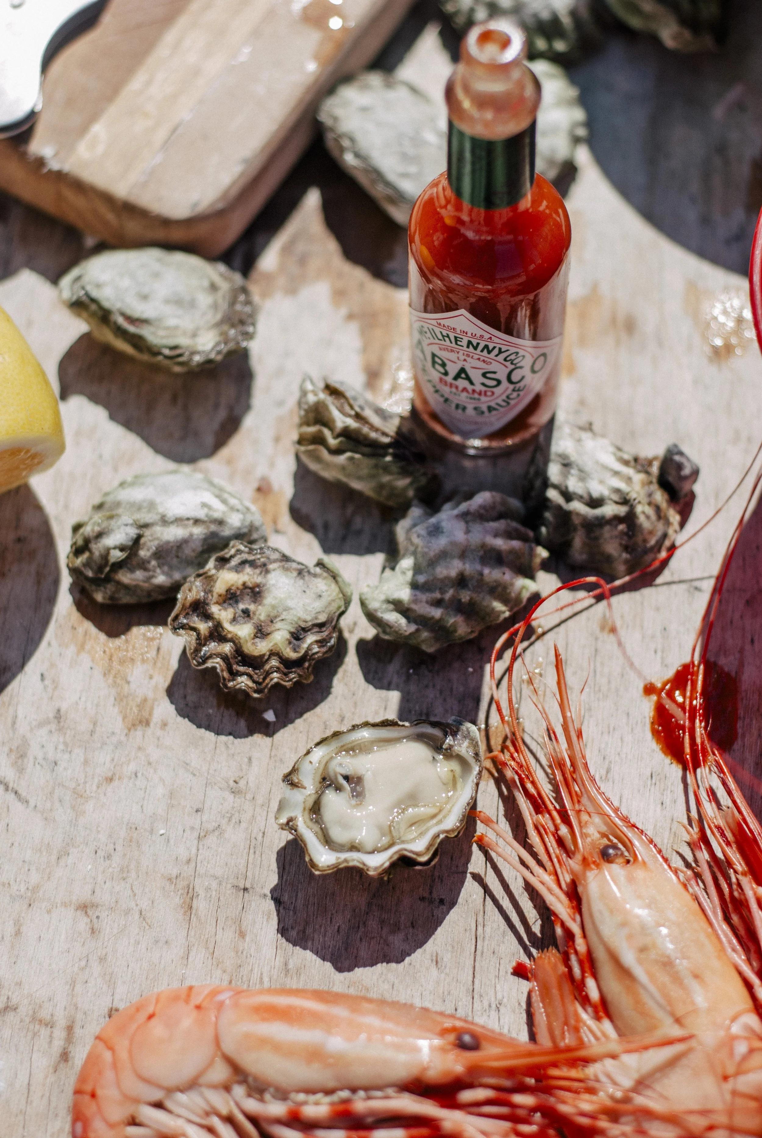 Fresh oysters on a wooden surface accompanied by a bottle of hot sauce and cooked spot prawns with orange antennae.