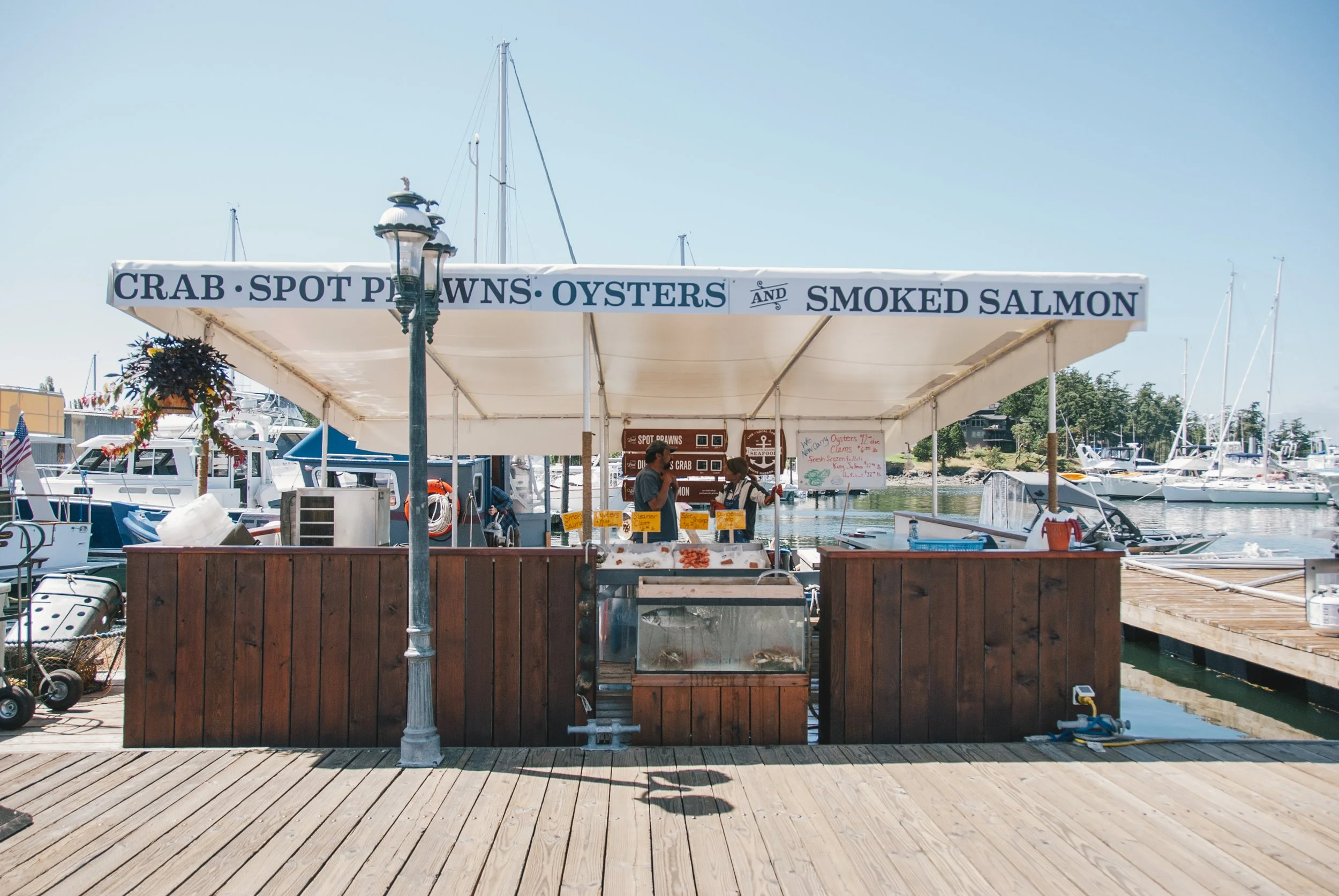 A marina with boats docked, a wooden stall selling seafood with a sign offering crab, spot prawns, oysters, and smoked salmon, and a person working at the seafood stall under a canopy.