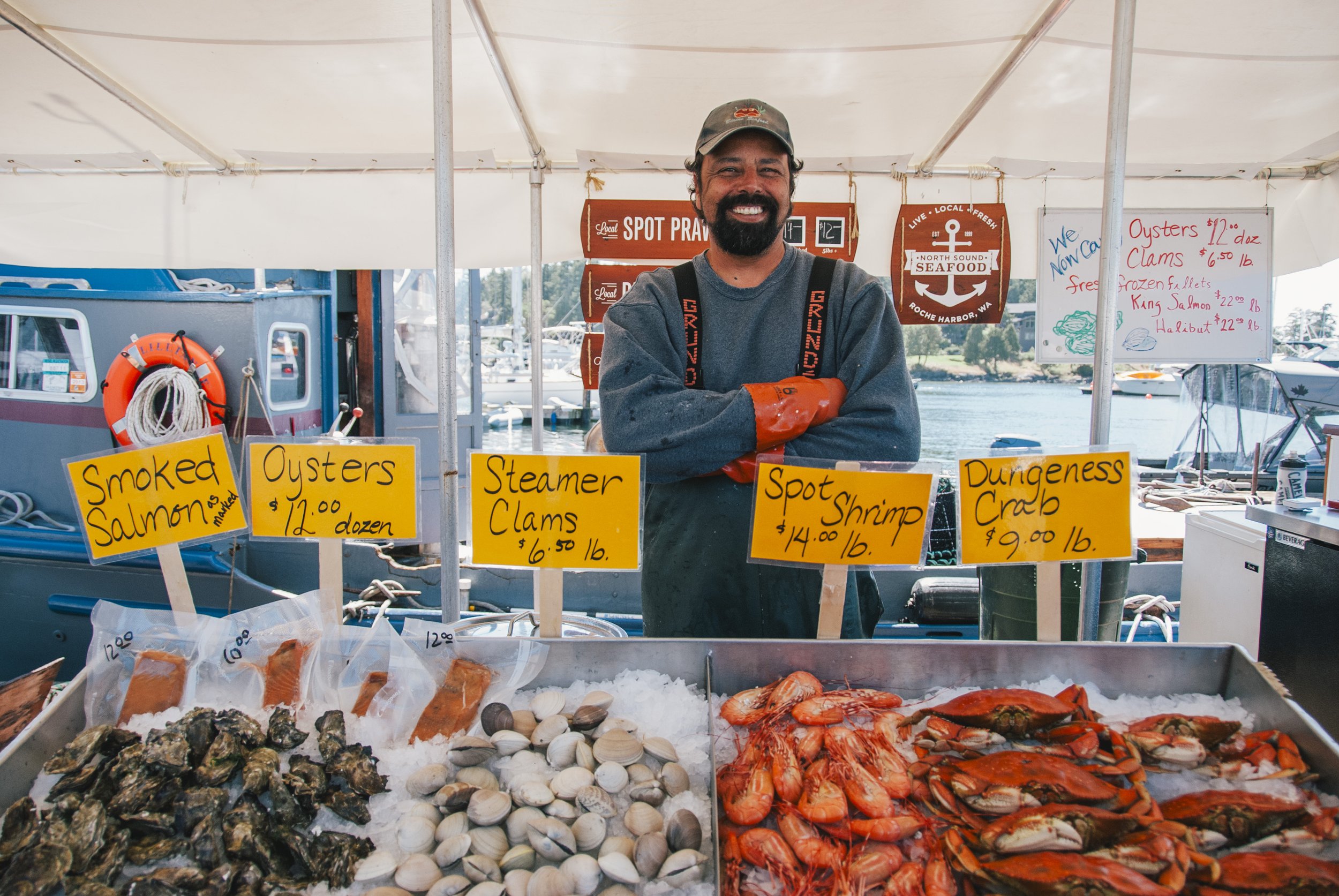 A smiling man standing behind a seafood display at a market with signs indicating prices for oysters, clams, shrimp, crab, and smoked salmon, with a boat and water in the background.