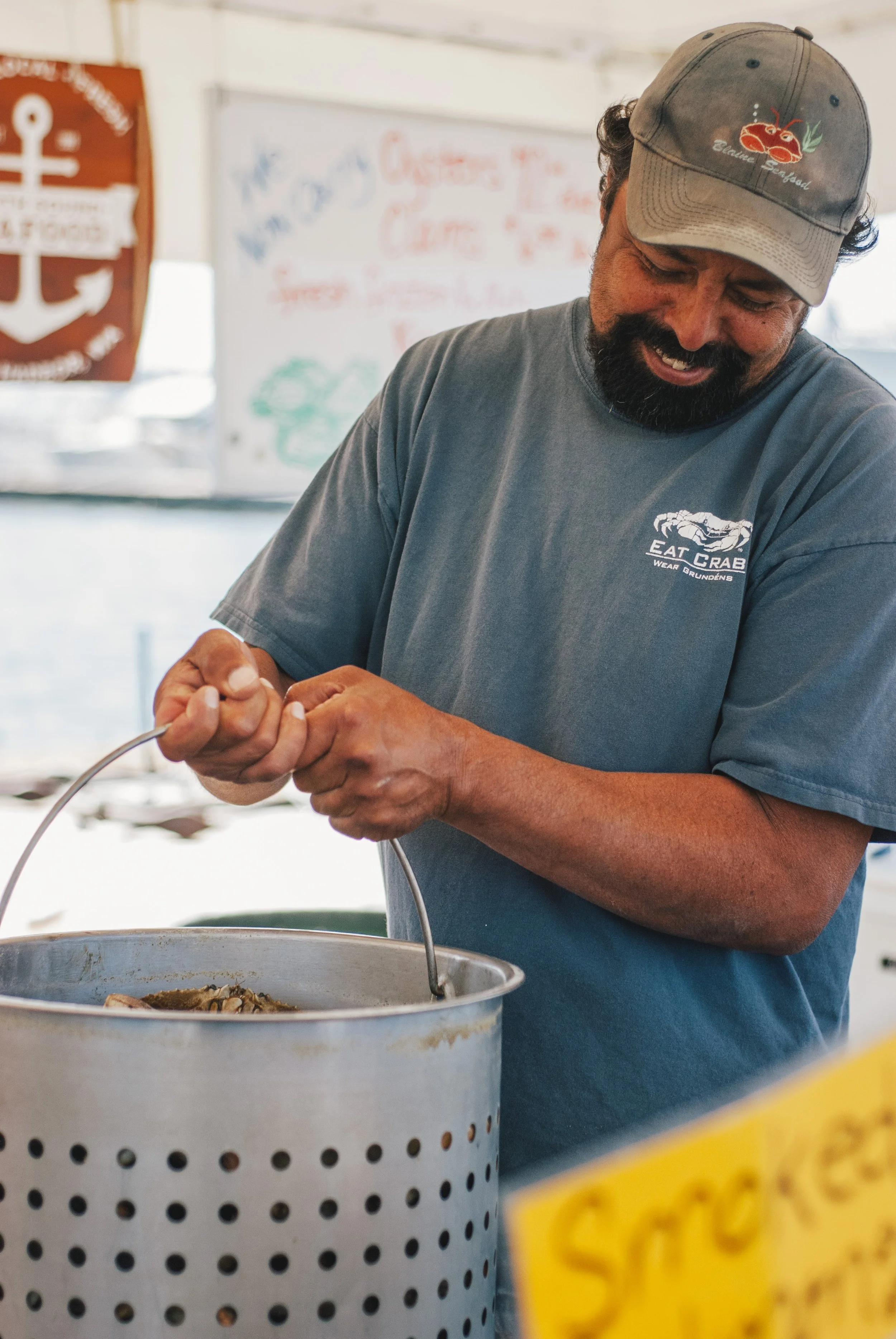 A man with a beard wearing a gray cap and a gray t-shirt labeled 'Eat Crab' is preparing seafood at a market stall. There is a basket in front of him and a sign with yellow and black writing partially visible.