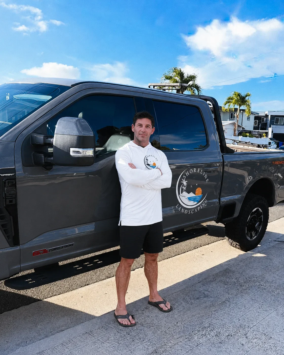 A man standing with arms crossed, wearing a white hoodie, black shorts, and flip-flops, standing next to a dark gray pickup truck with a lake and sunset logo on the door that reads 'Shoreline Landscaping', on a sunny day with a blue sky, palm trees, and houses in the background.