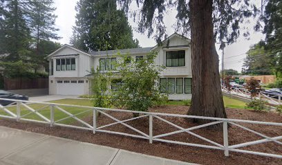 A two-story house with a white exterior, surrounded by tall trees and a white wooden fence in the front yard.