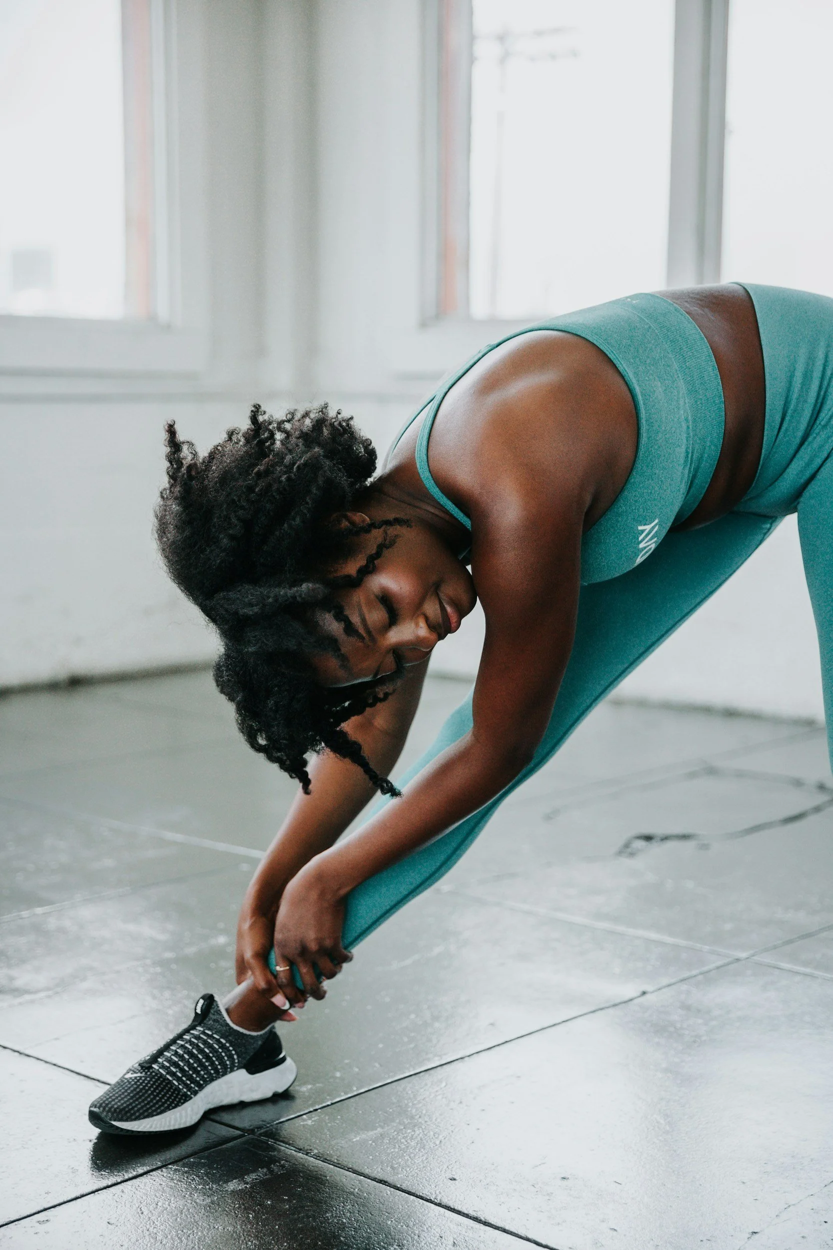 A woman in a teal sports bra and matching leggings doing a forward bend stretch in a brightly lit room with large windows, black athletic shoes, and a gray tiled floor.