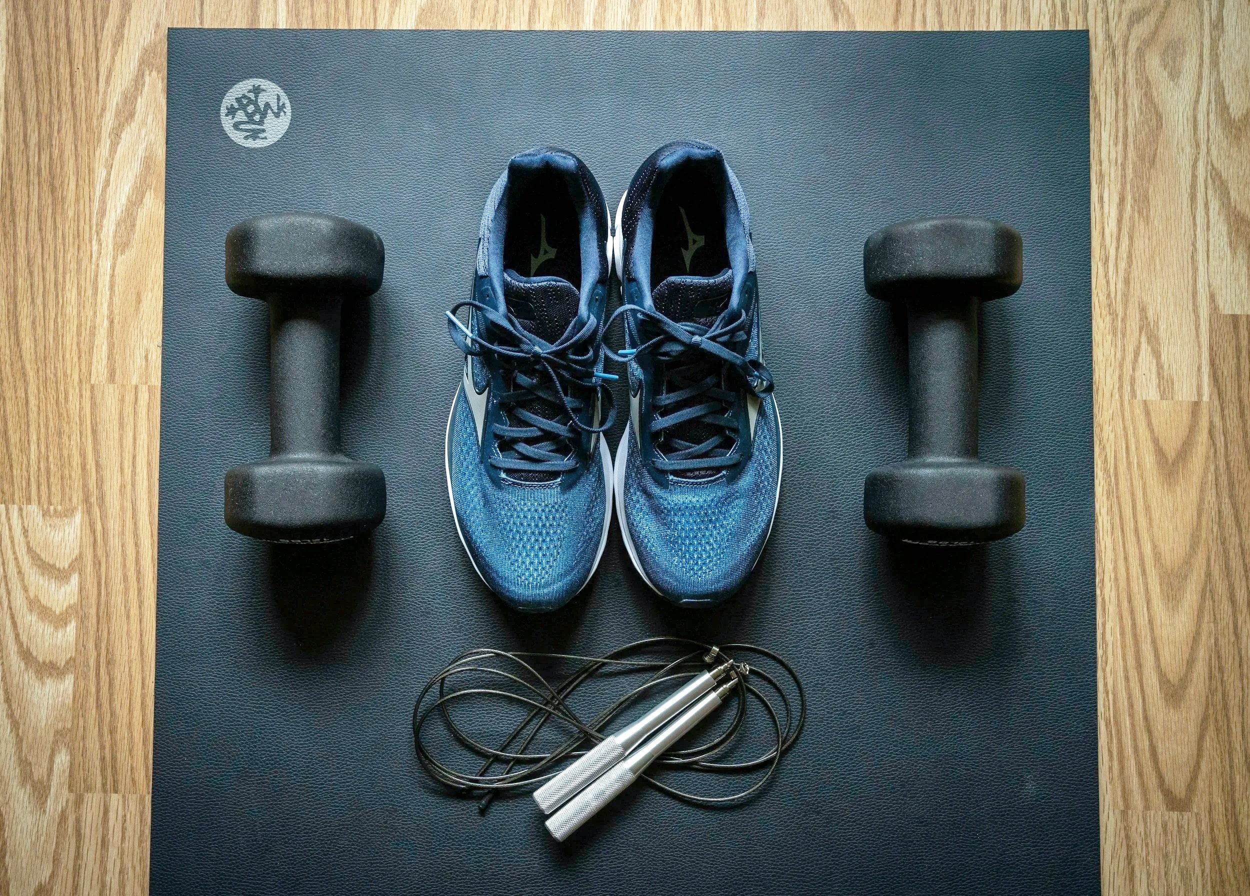 a pair of blue running shoes on a black yoga mat next to two black dumbbells and a jump rope, all on a wooden floor
